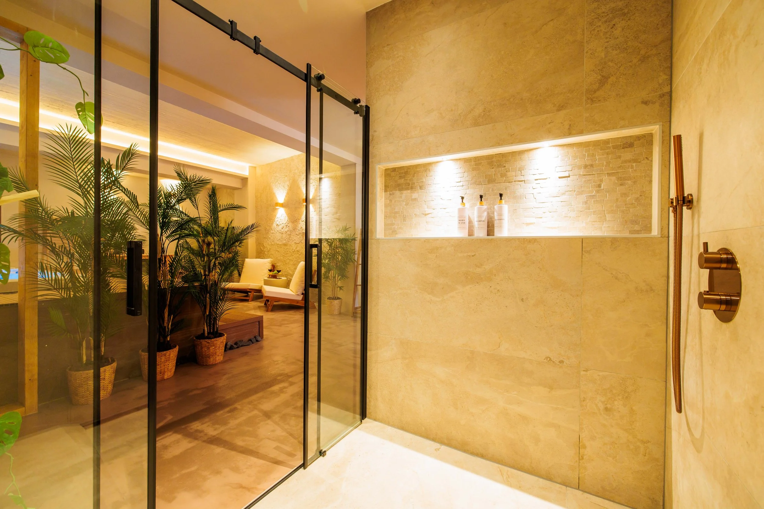 Luxurious bathroom shower area with beige stone walls, a niche with three bottles, and a glass door leading to a cozy room with plants and seating.