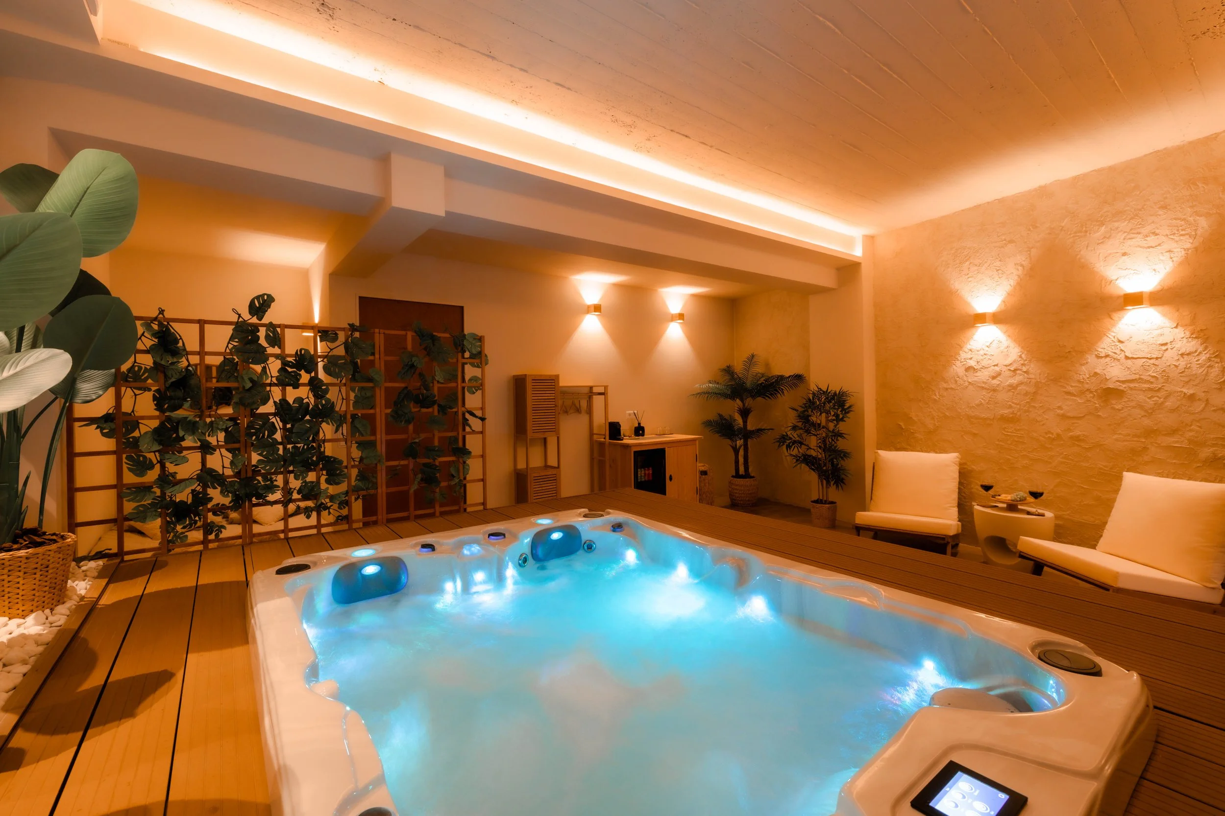 Indoor spa room with a lit whirlpool tub, seating area with white chairs, plants, and warm lighting.