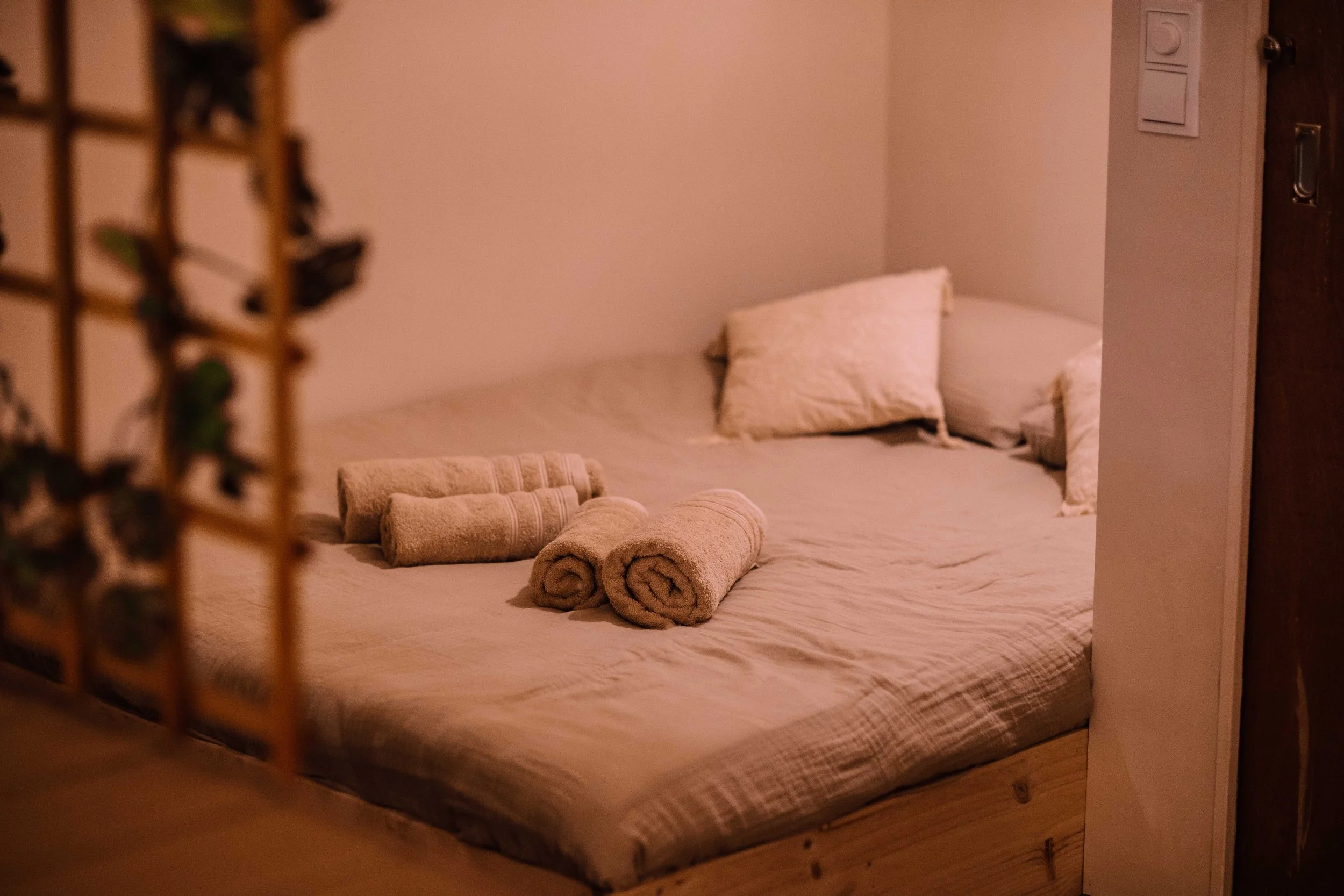 A neatly made bed with two rolled towels and two pillows on a wooden bed frame, in a softly lit room.
