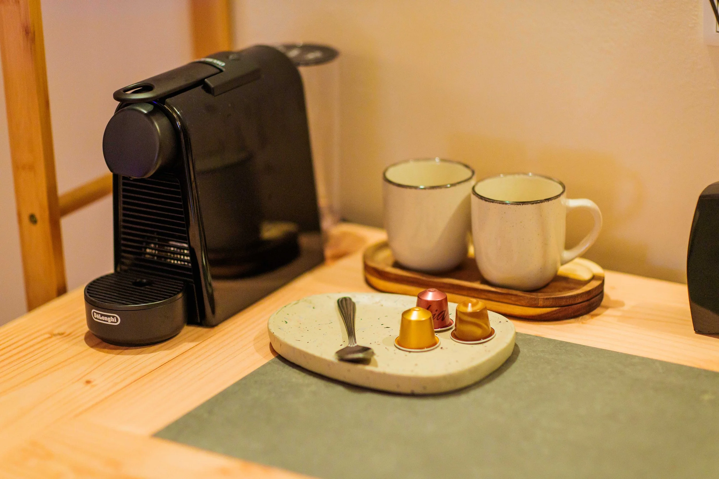 Coffee station with a black coffee machine, two white ceramic mugs on a wooden tray, a green speckled plate with a small spoon and colorful coffee capsules on a wooden table.