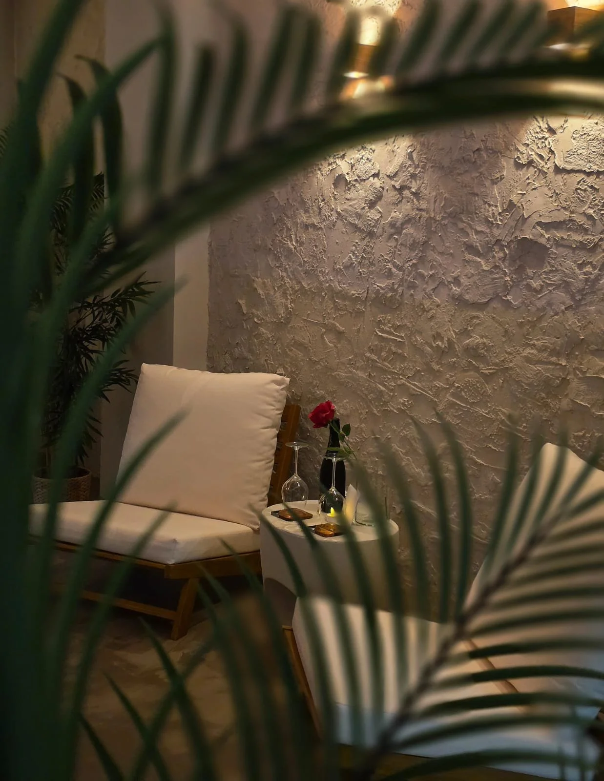 A cozy indoor seating area with a white cushioned lounge chair, a small round side table with a single red rose in a black vase, a glass with a stem, and a wine opener, framed by green palm leaves in the foreground. The background features a textured off-white wall.
