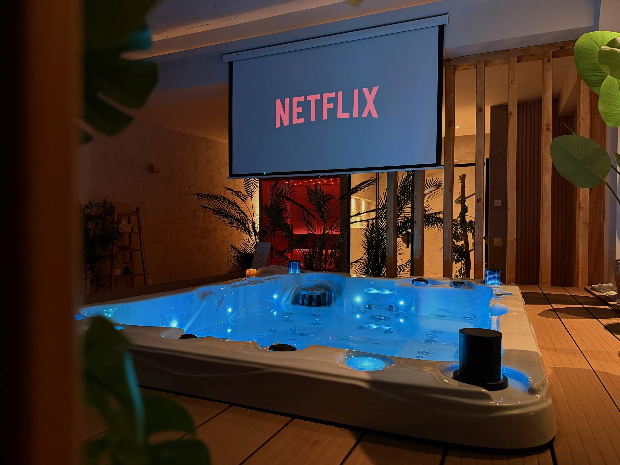 Indoor Jacuzzi with blue lights and a large Netflix screen above it, surrounded by plants and warm lighting.