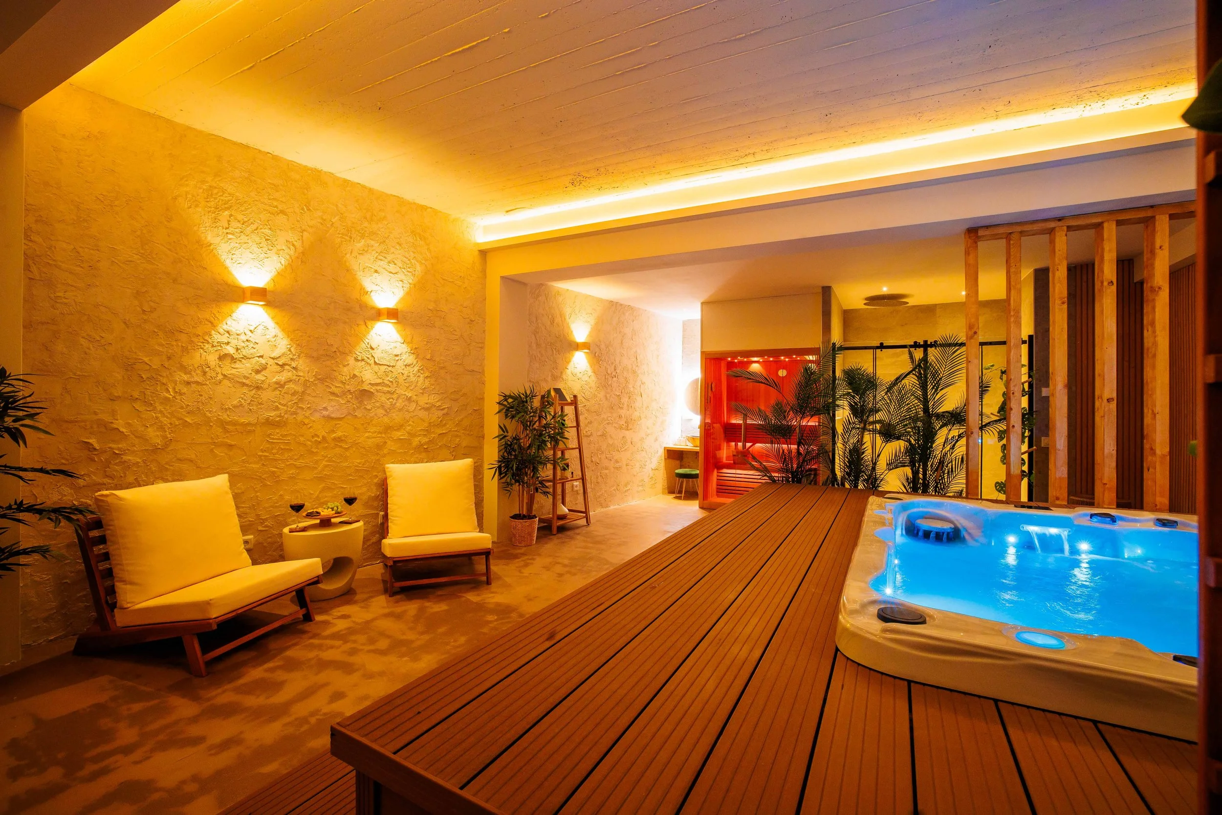 Indoor private spa room with a jacuzzi with blue lighting, wooden flooring, beige lounge chairs with yellow cushions, potted plants, soft warm lighting on textured walls, and a wooden partition with plants in the background.