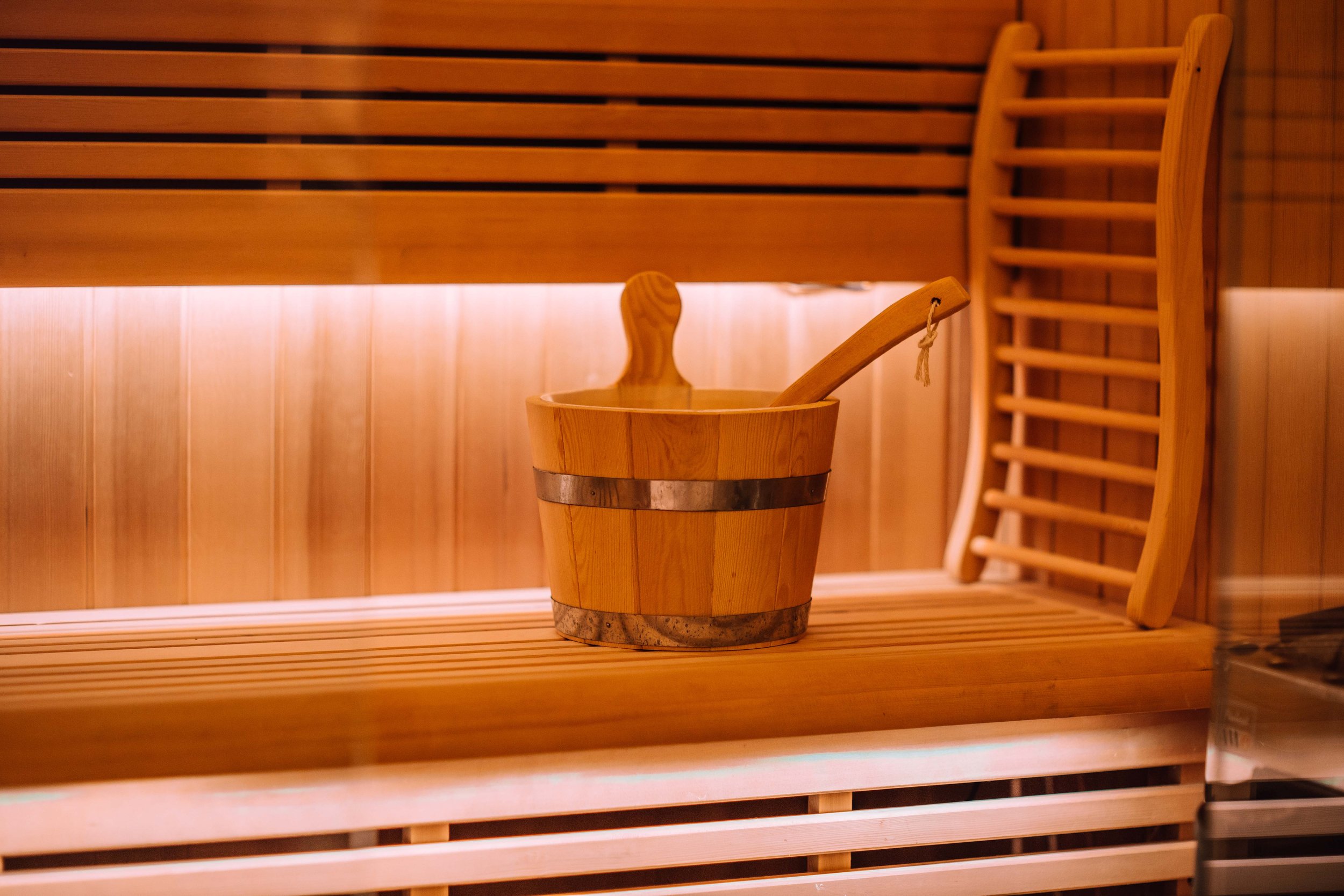 Wooden sauna interior with a bucket and ladle on a bench, and a wooden chair in the background.