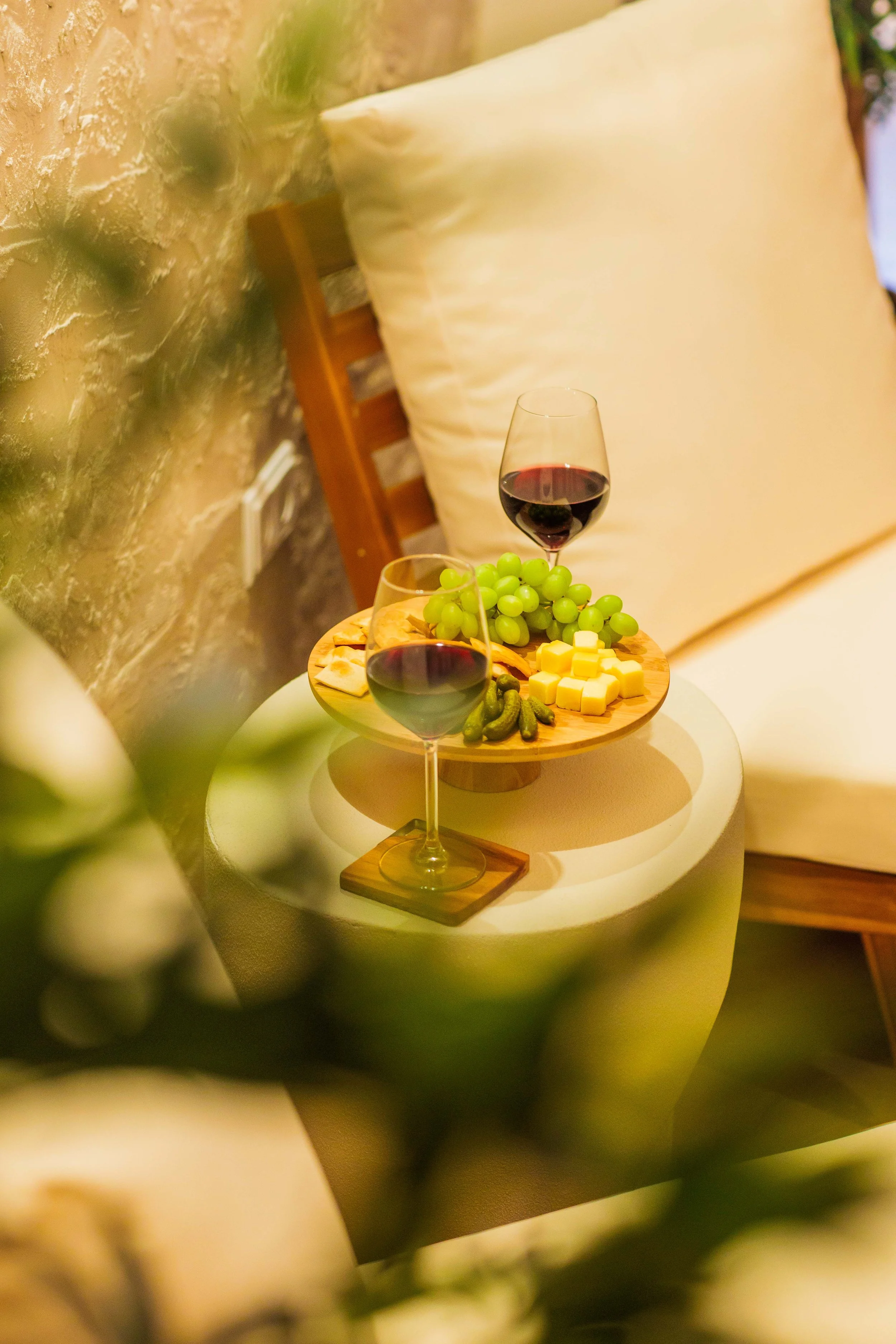 A cozy bedroom scene with a small round table holding two glasses of red wine, a wooden cheese and fruit platter with green grapes, cheese cubes, crackers, and pickles. A bed with a cream pillow and a wooden headboard are visible in the background.