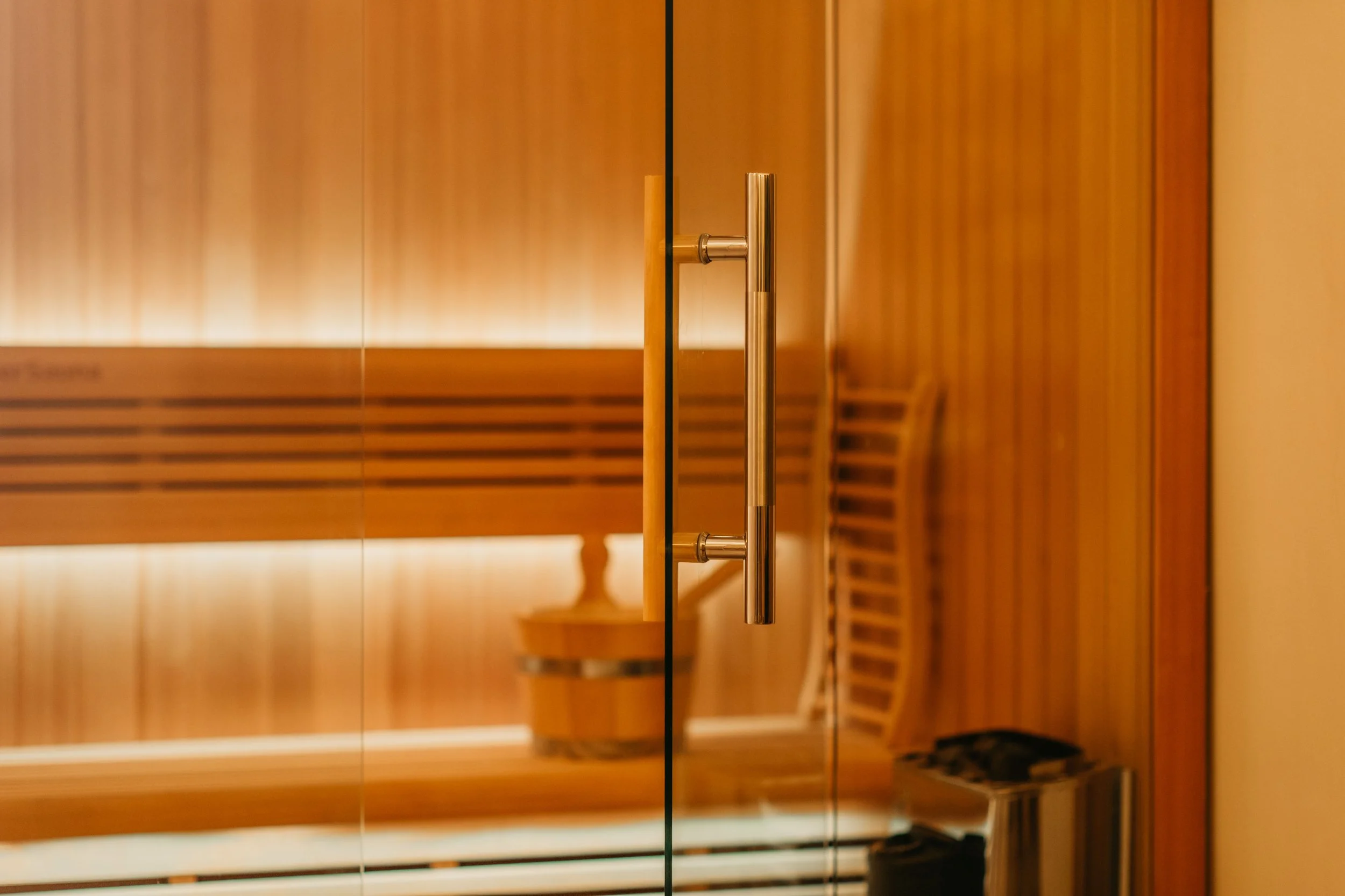 Close-up of a glass door handle inside a sauna with wooden walls and a wooden chair reflected in the glass.
