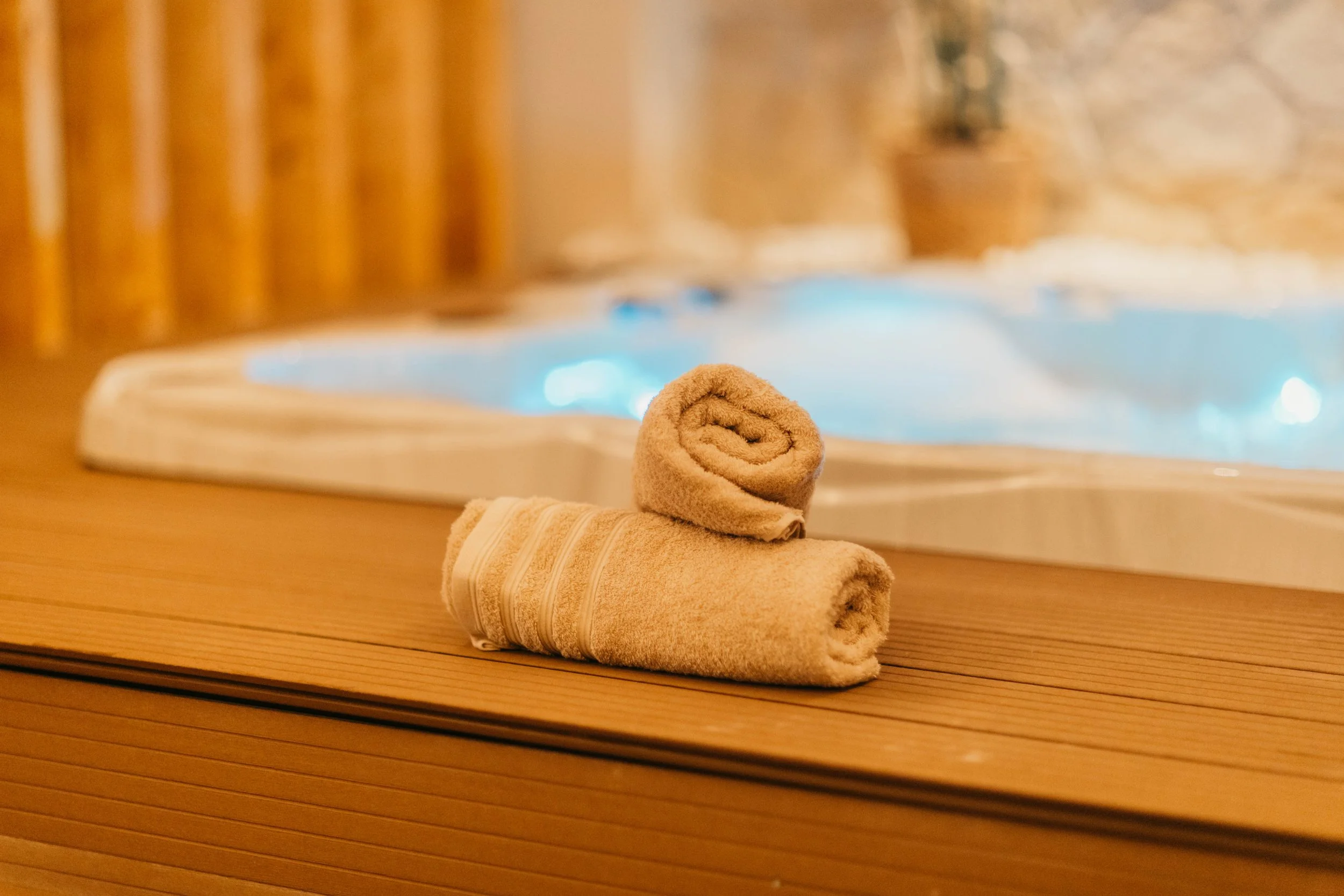 Two rolled towels, one beige and one cream, resting on a wooden edge near a hot tub with blue water.