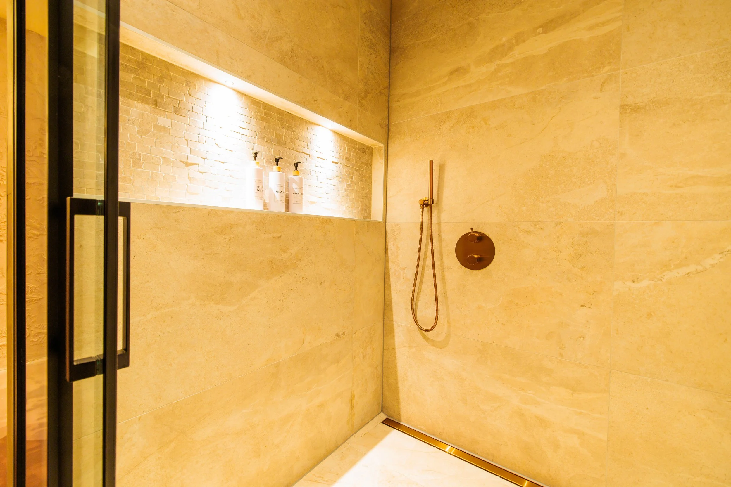 A minimalist walk-in shower with beige marble walls and a built-in niche with lighting, holding three white bottles. The shower features a black fixture with a handheld showerhead and a circular control panel on the wall.