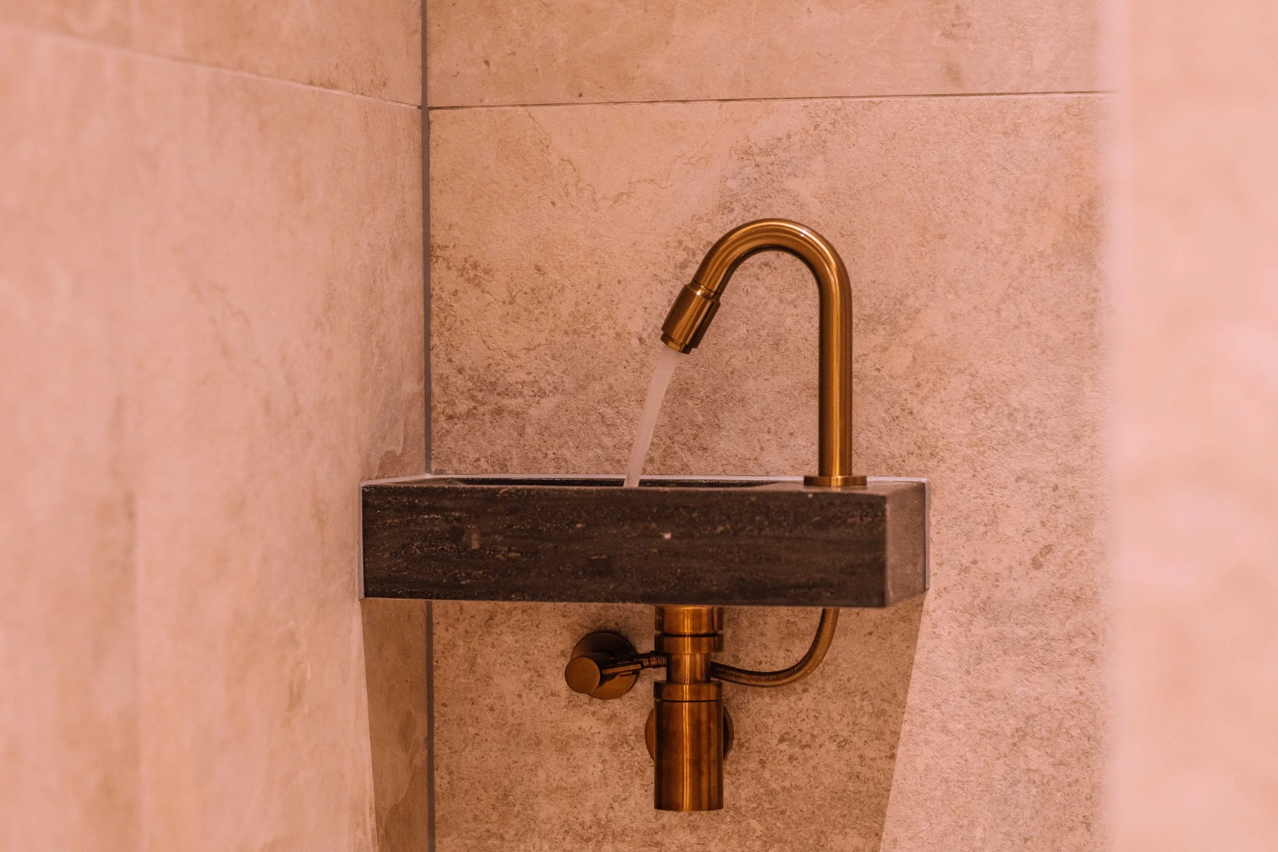 A gold faucet with a stream of water over a dark stone sink, mounted on a beige tiled wall.
