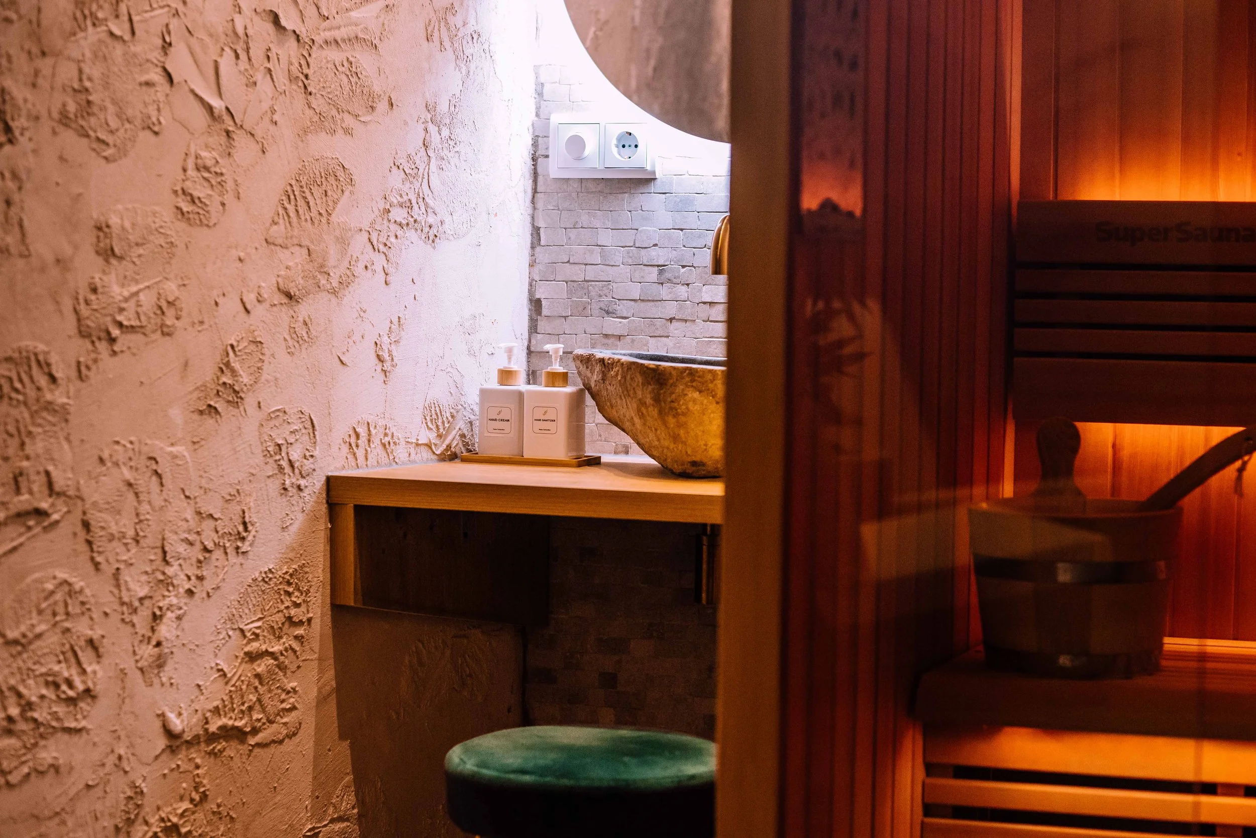 A cozy spa room with a stone sink, bottles of hand soap on a wooden counter, and a steam room with a wooden door labeled 'SuperSauna'.