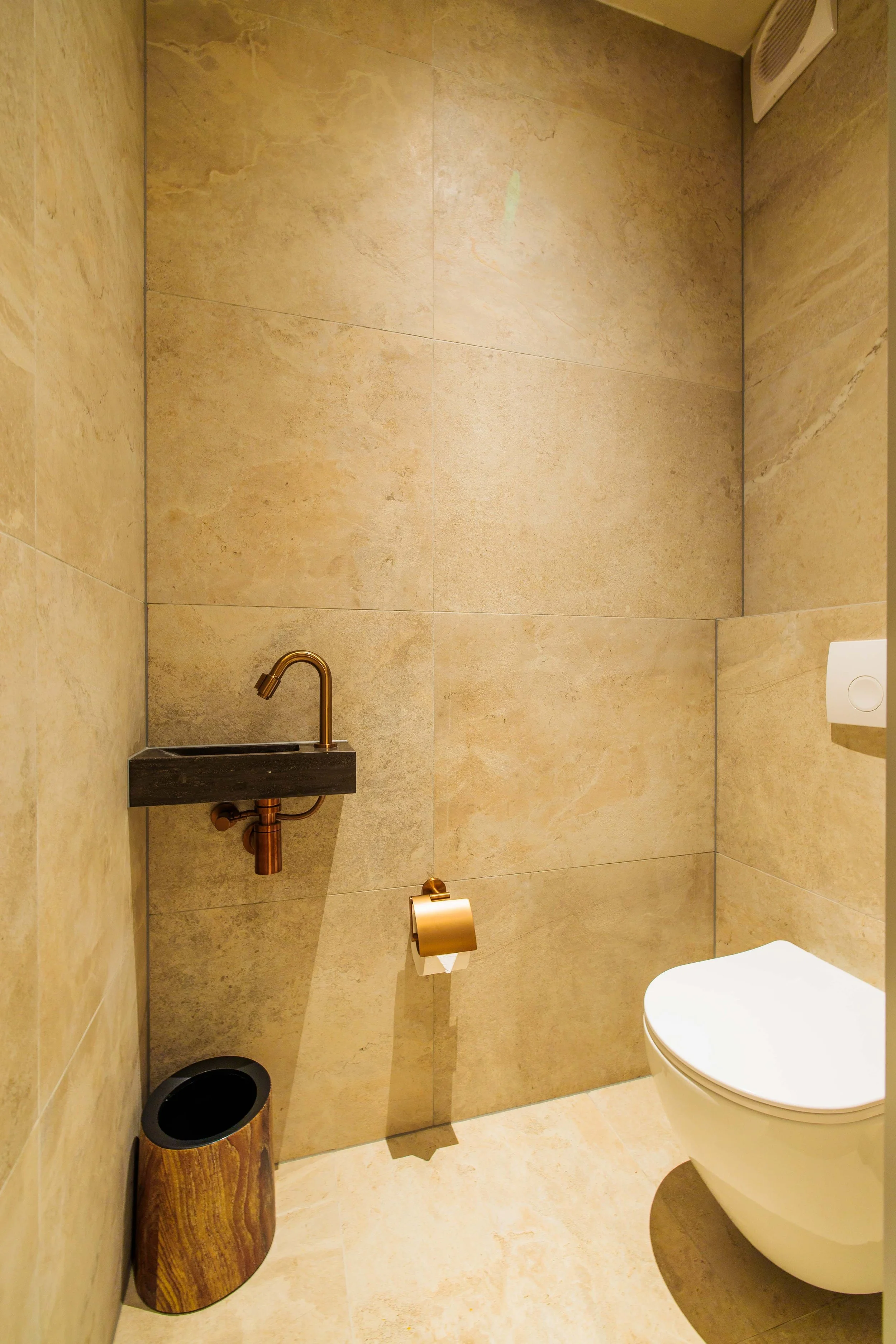 A modern minimalist bathroom with beige tiled walls and floor, a white wall-mounted toilet, a small black sink with a copper faucet, a coin-operated toilet paper dispenser, and a small wooden trash bin.