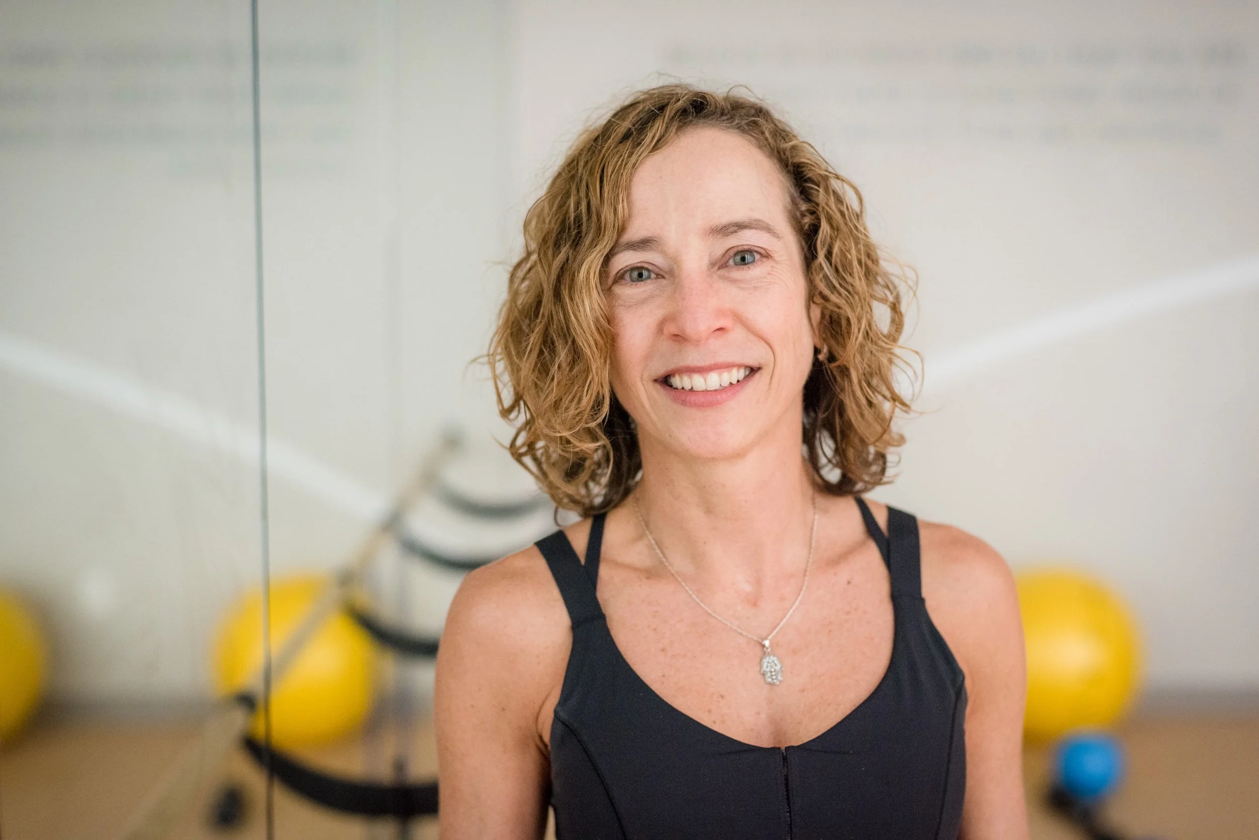 A smiling woman with curly blonde hair wearing a black tank top and a silver necklace, standing in a gym with yellow and blue exercise balls in the background.