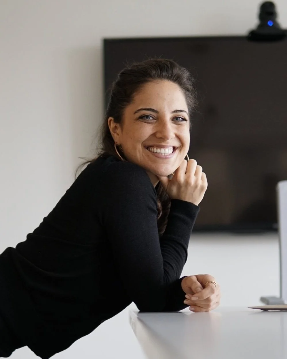 A woman with dark hair smiling, sitting at a table with her chin resting on her hand, in an office setting with a large monitor behind her.