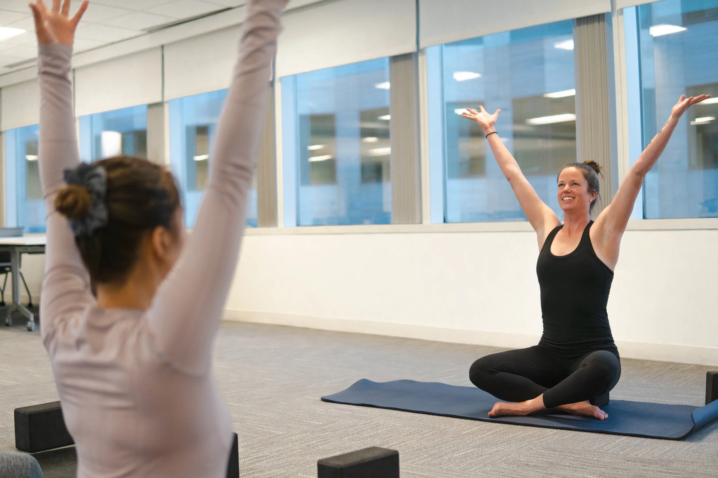Two women participating in a yoga class indoors, sitting on yoga mats with arms raised in a celebratory pose, windows in the background.