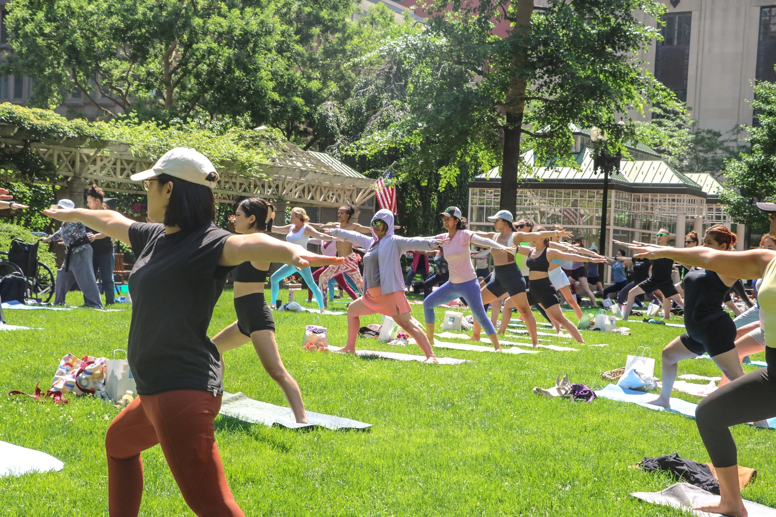 Group of people participating in a yoga class outdoors in a park, practicing the Warrior pose on yoga mats.