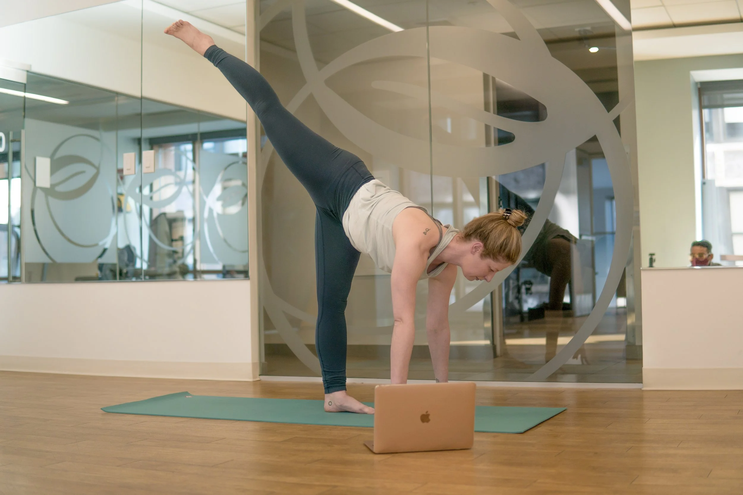 Woman practicing yoga in a gym, performing a forward bend with one leg extended upward, in front of a glass wall with a circular design, a yoga mat, and a laptop nearby.