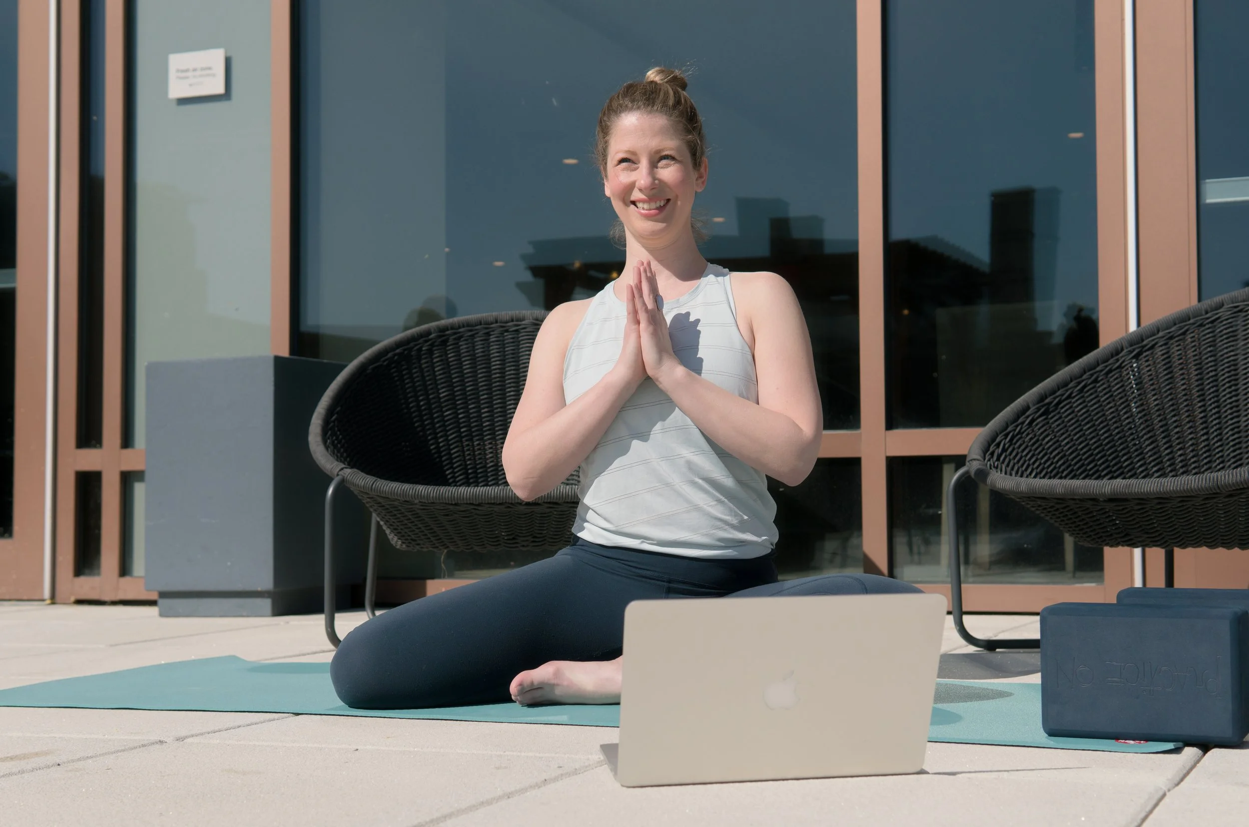 Woman practicing yoga outdoors on a patio, sitting on a yoga mat with hands together in prayer pose, laptop and workout equipment nearby.