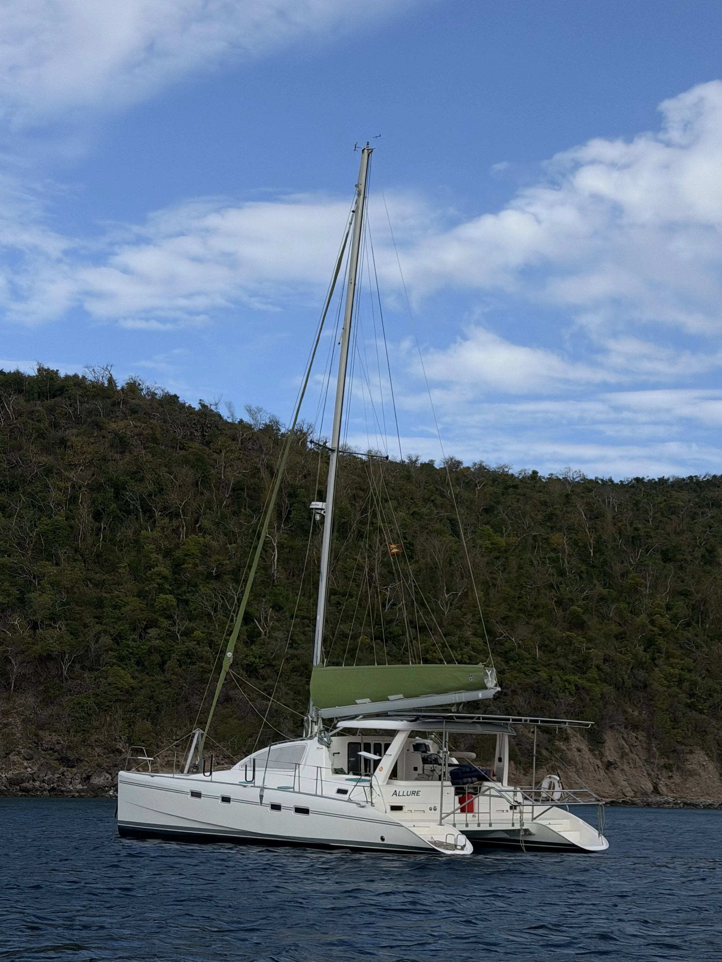 A white sailing catamaran named 'ALLURE' with a green canopy, anchored near a hill covered with trees, under a partly cloudy blue sky.