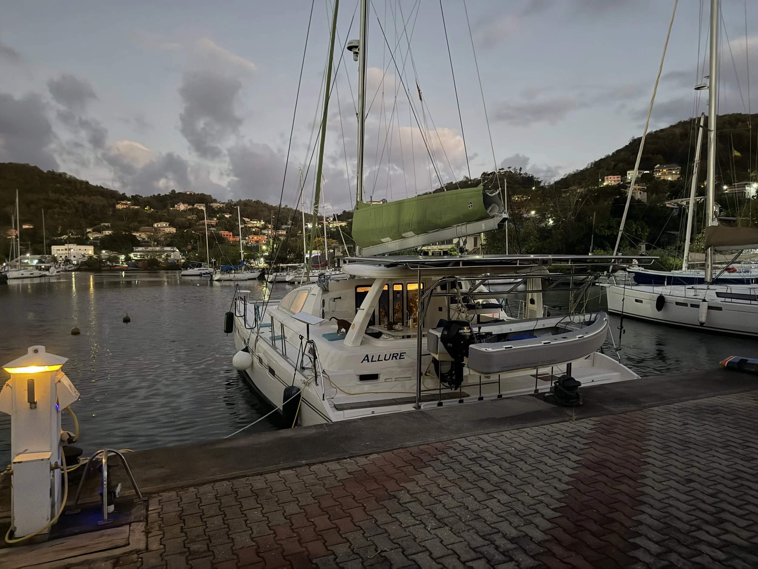 A marina at dusk with boats docked, including a white sailing catamaran named 'Allure,' electric power pedestals, a hillside with houses, and cloudy skies.