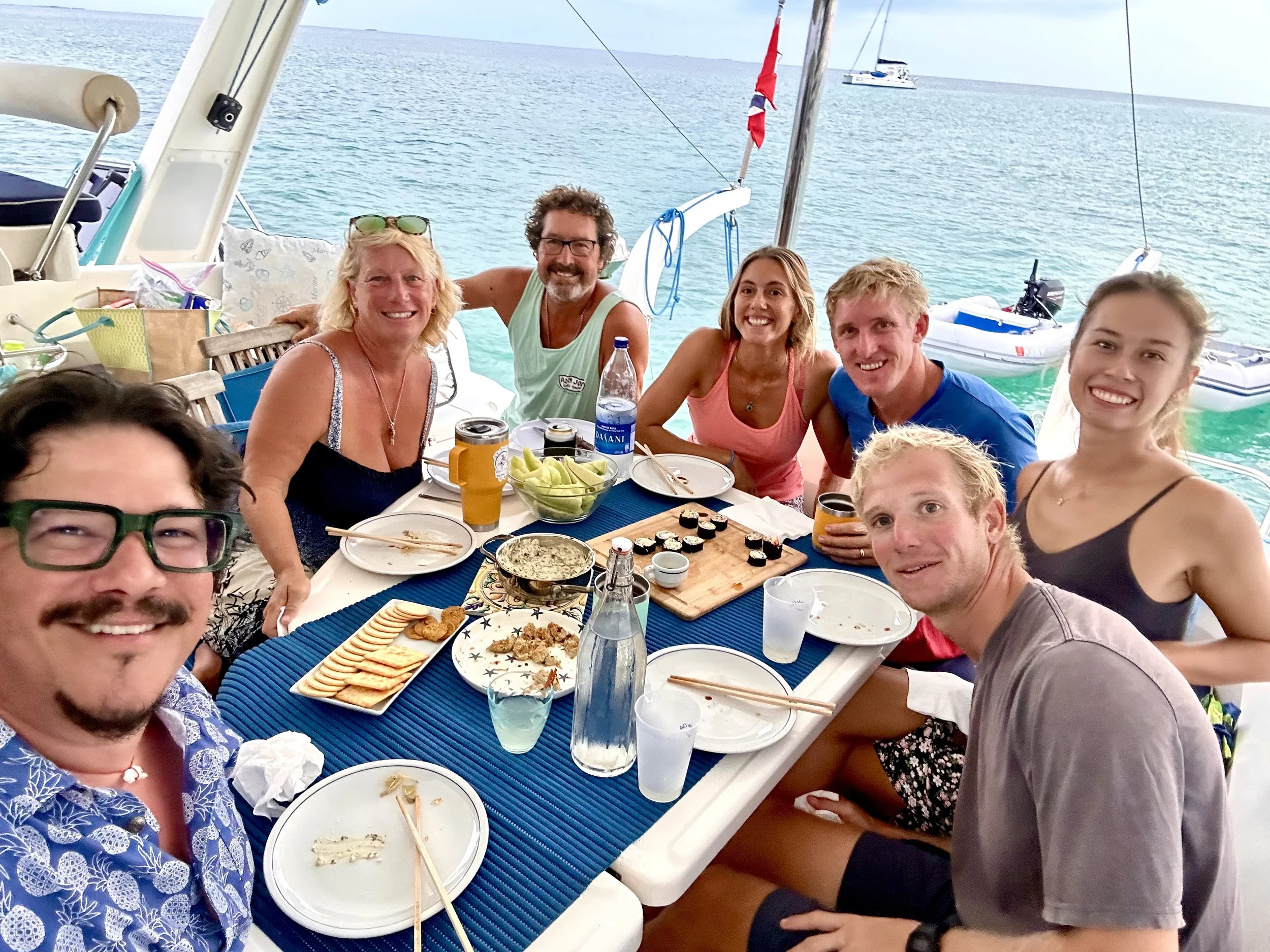 Captain Joe and group of friends enjoying a meal aboard Allure over water, with boats on anchor and the open sea visible in the background, and a table set with sushi, drinks, and plates.