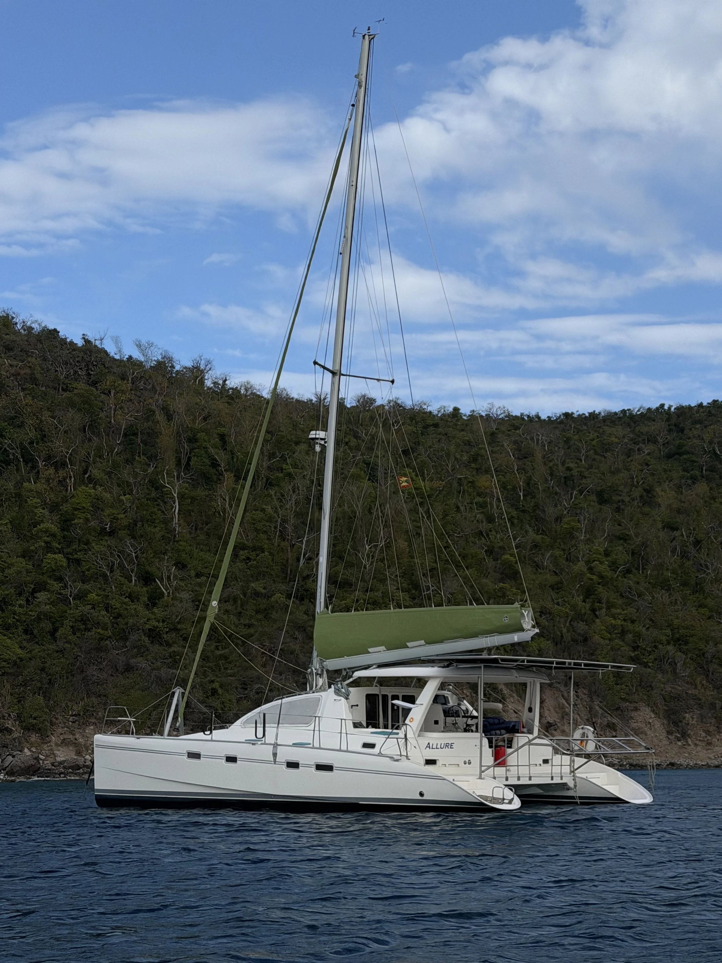 A white sailing catamaran named 'ALLURE' anchored near a shoreline with a green mountain and partly cloudy blue sky in the background.