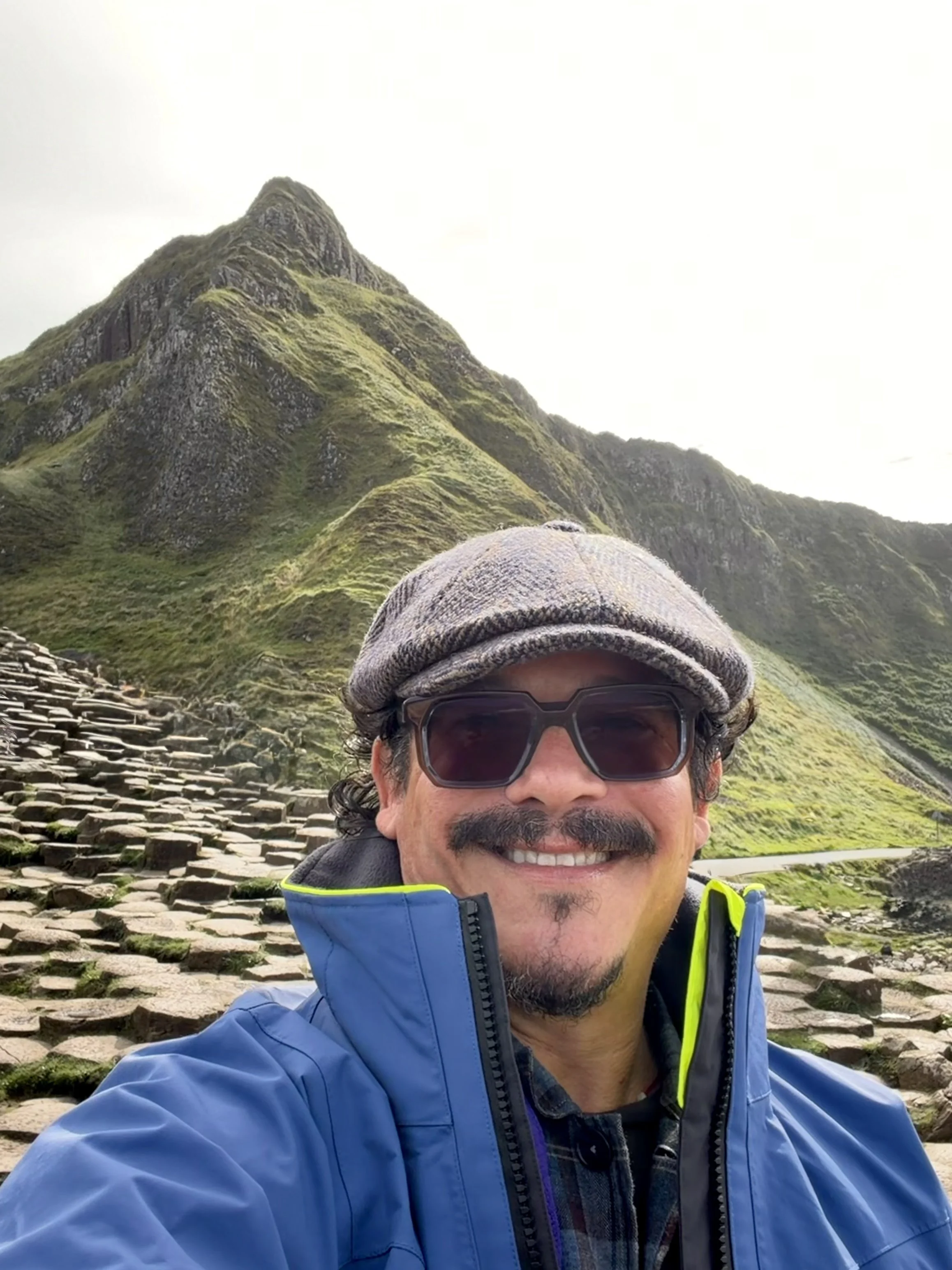 Captain Joe wearing a gray baker's cap, sunglasses, and a blue sailing jacket taking a selfie outdoors with lush green mountains and stone steps in the background at Giant's Causeway in Ireland.