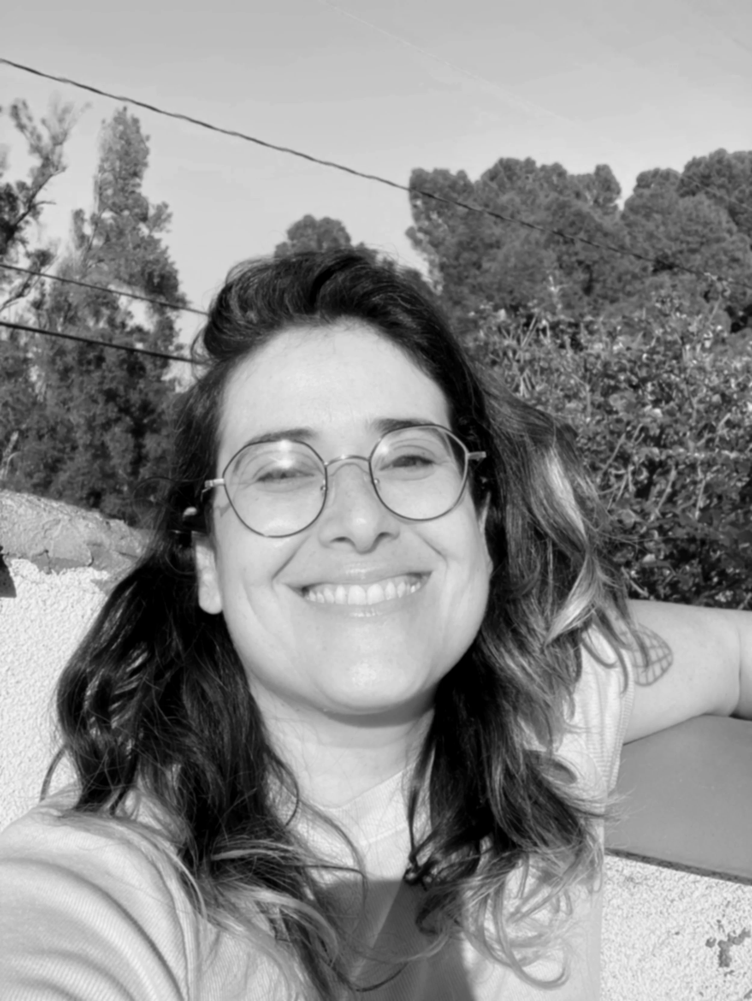A woman with dark, wavy hair, glasses, and a big smile, outdoors on a sunny day with trees and power lines in the background.