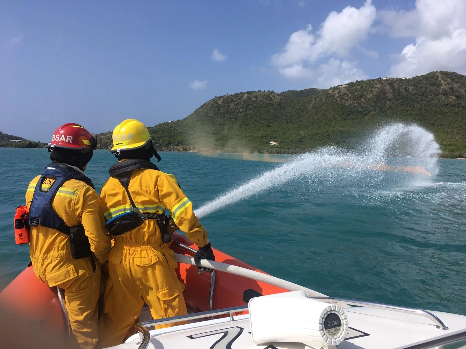Two lifeguards in yellow uniforms and helmets on a rescue boat, spraying water during a rescue operation near an island with hilly terrain.