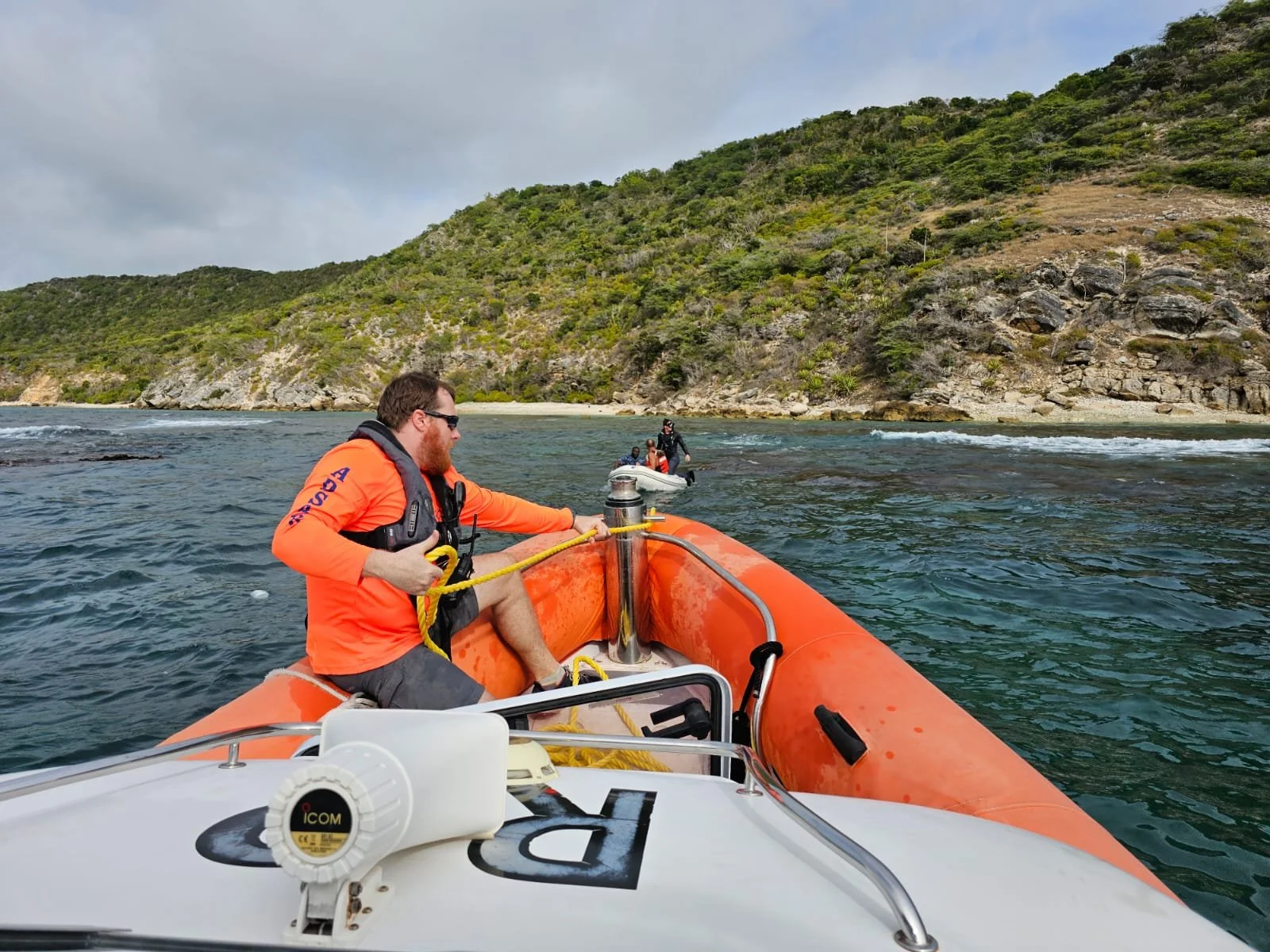 A person in an orange life jacket and black shirt sitting on an orange inflatable boat on the water, holding a yellow rope. Two other people are visible in the distance, also on inflatable boats, near a green hill with rocky terrain.