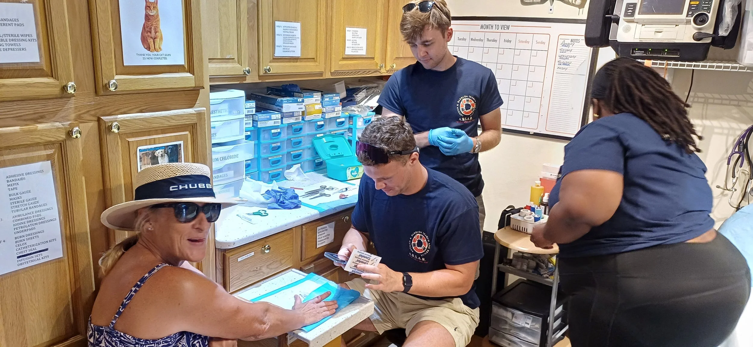Four people, three men and one woman, are gathered in a medical supply room. The woman is seated with her arm extended, receiving medical treatment. The man seated is looking at pamphlets or medical instructions. The other two people are standing, wearing gloves, and attending to the medical supplies and equipment on the counter. The room has wooden cabinets, shelves with medical supplies, and a whiteboard with notes.