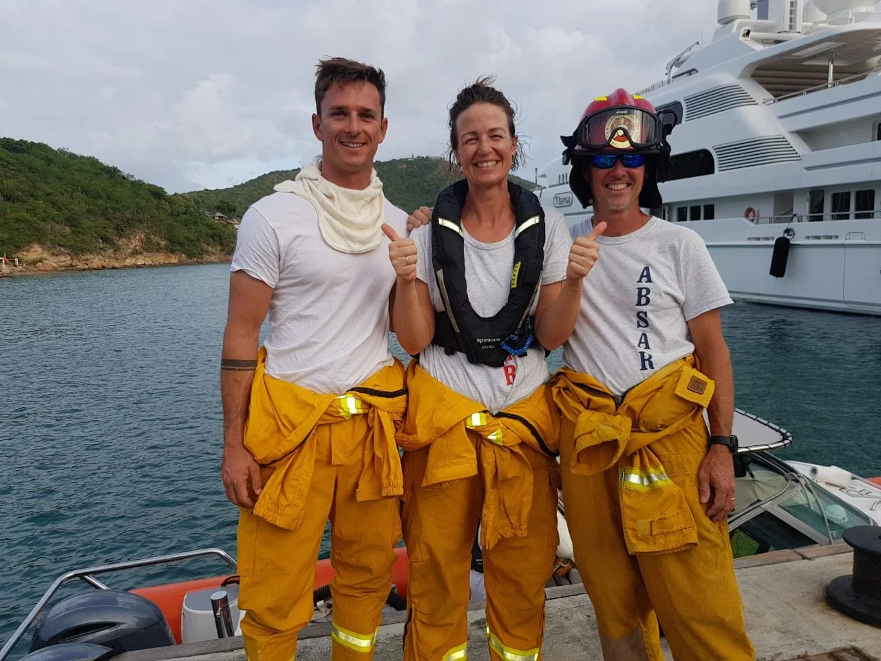 Three people in firefighter gear standing on a dock with boats and a hillside in the background. The person on the right is wearing a firefighter helmet and sunglasses, the middle person is smiling with thumbs up, and the left person is smiling with a cloth around their neck.