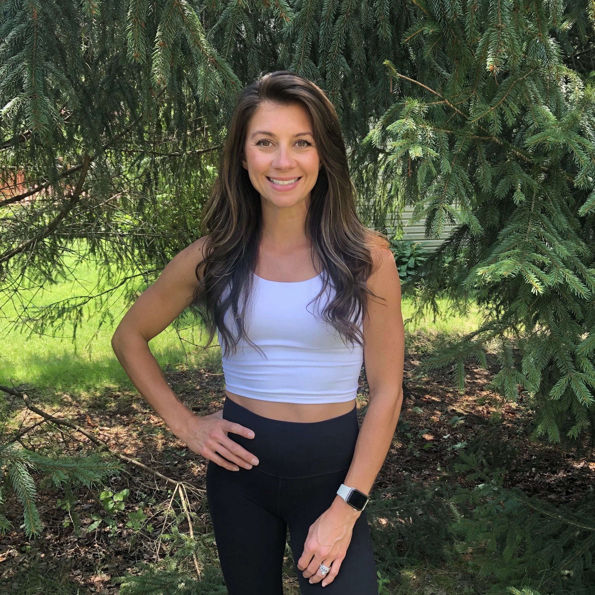 A woman standing outdoors in front of green trees, smiling and wearing a white crop top and black pants.