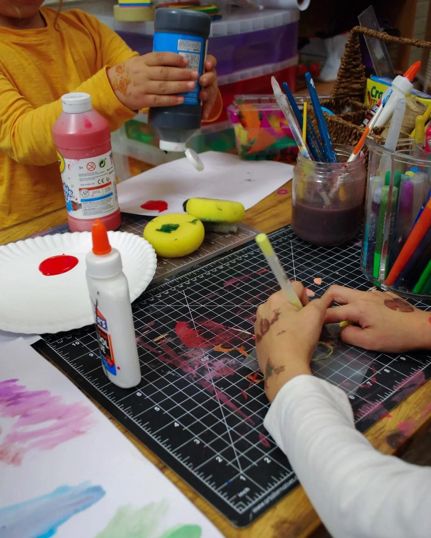 Children engaging in arts and crafts, using paints, glue, and brushes on a black cutting mat with colorful markers and crafting supplies on a wooden table.