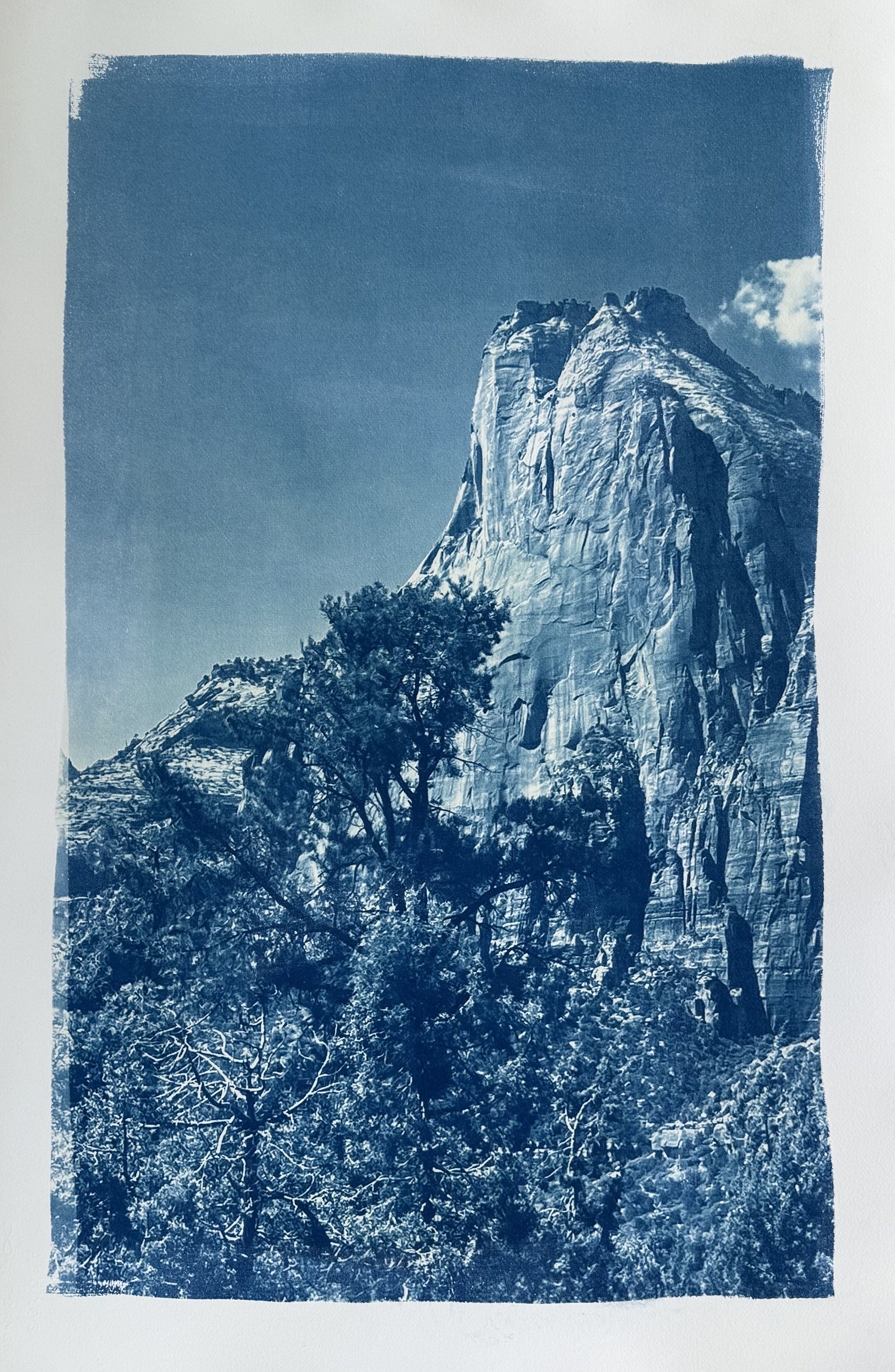 A monochromatic blue-toned image of a mountain with steep cliffs, a few trees in the foreground, and a partly cloudy sky.