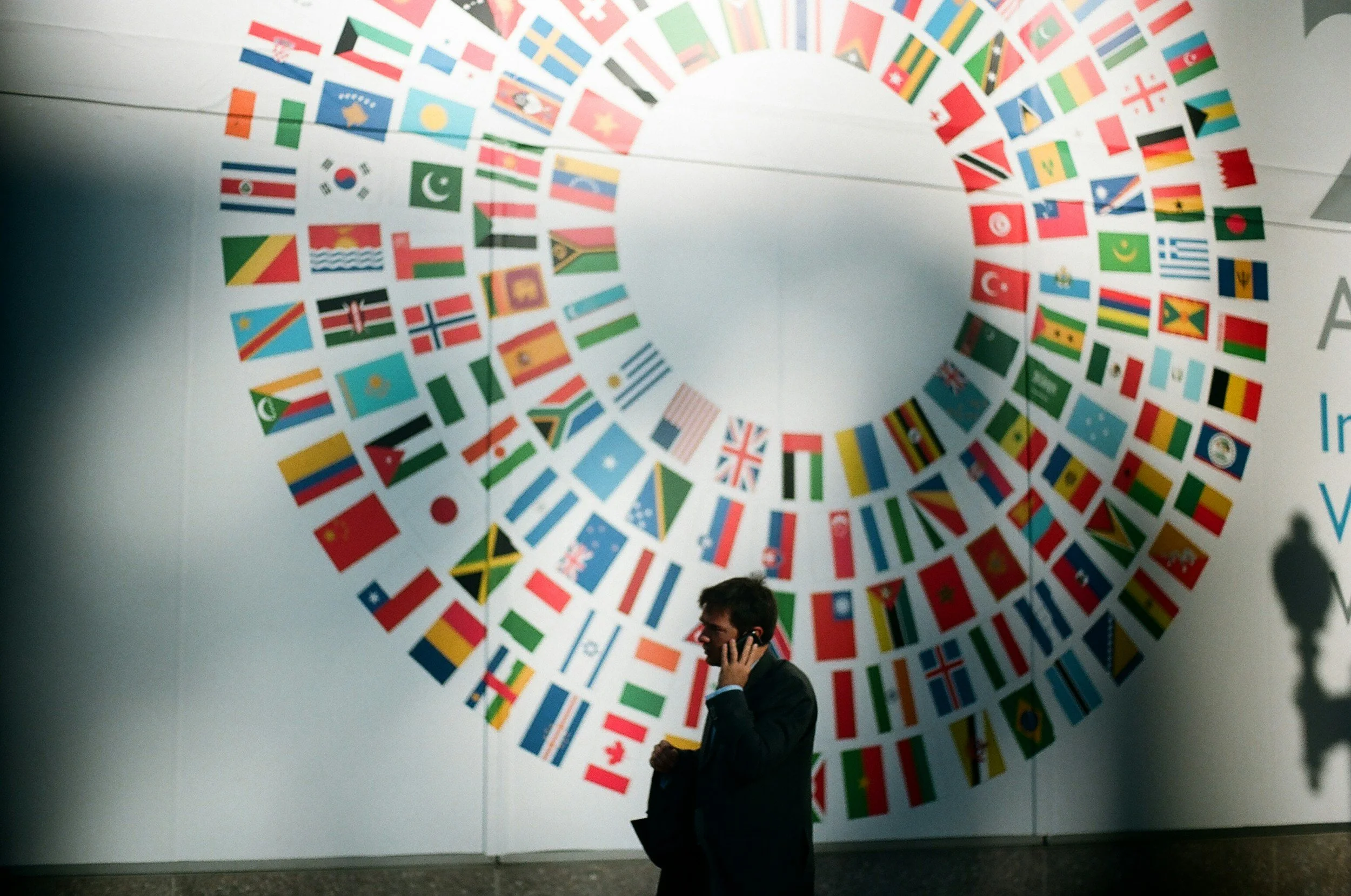 A large circular display of numerous international flags on a wall, with a man in a suit talking on a cell phone in the foreground.