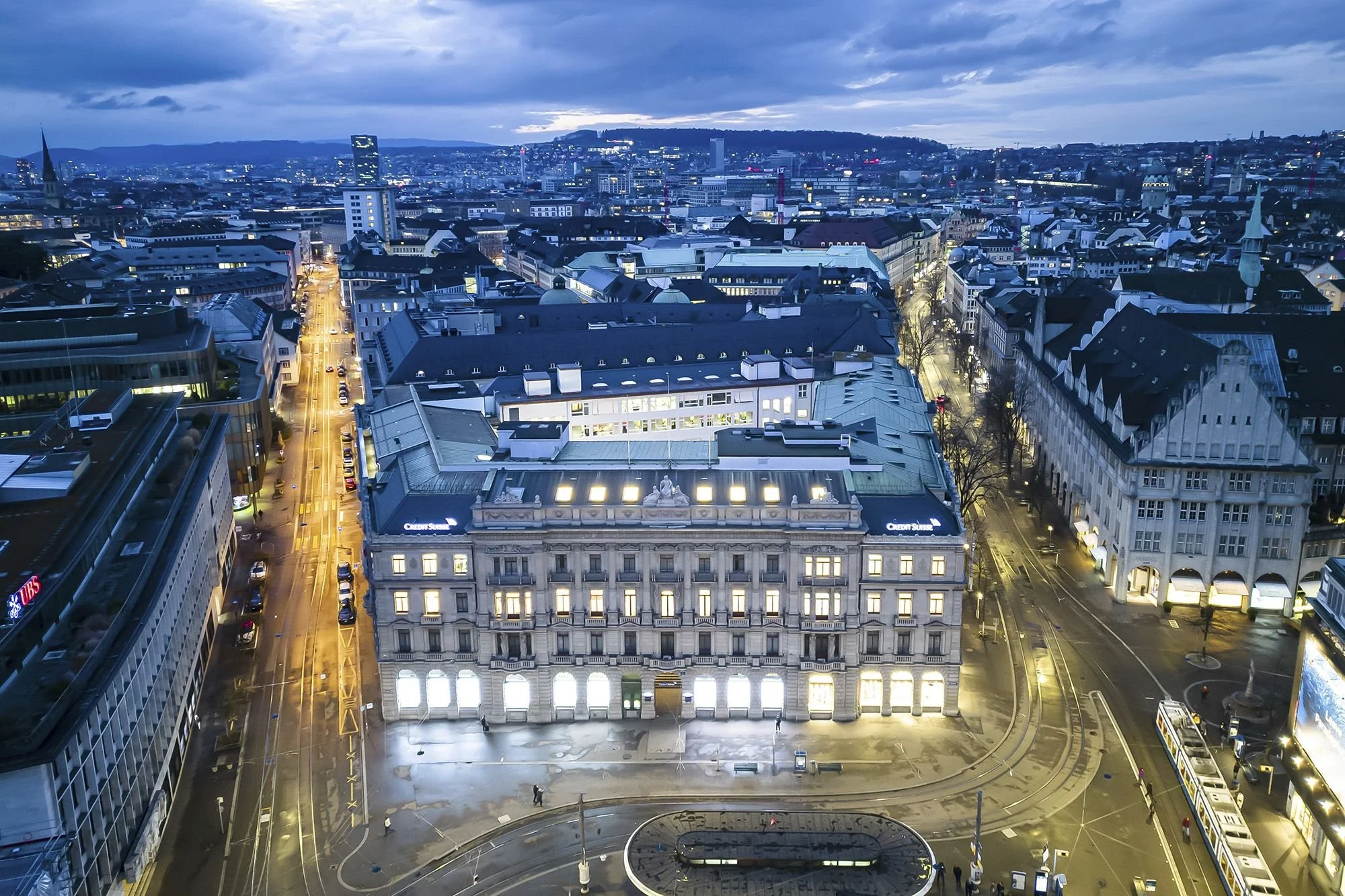 Aerial view of a cityscape at dusk with illuminated buildings and streets, including a prominent historic building in the foreground.