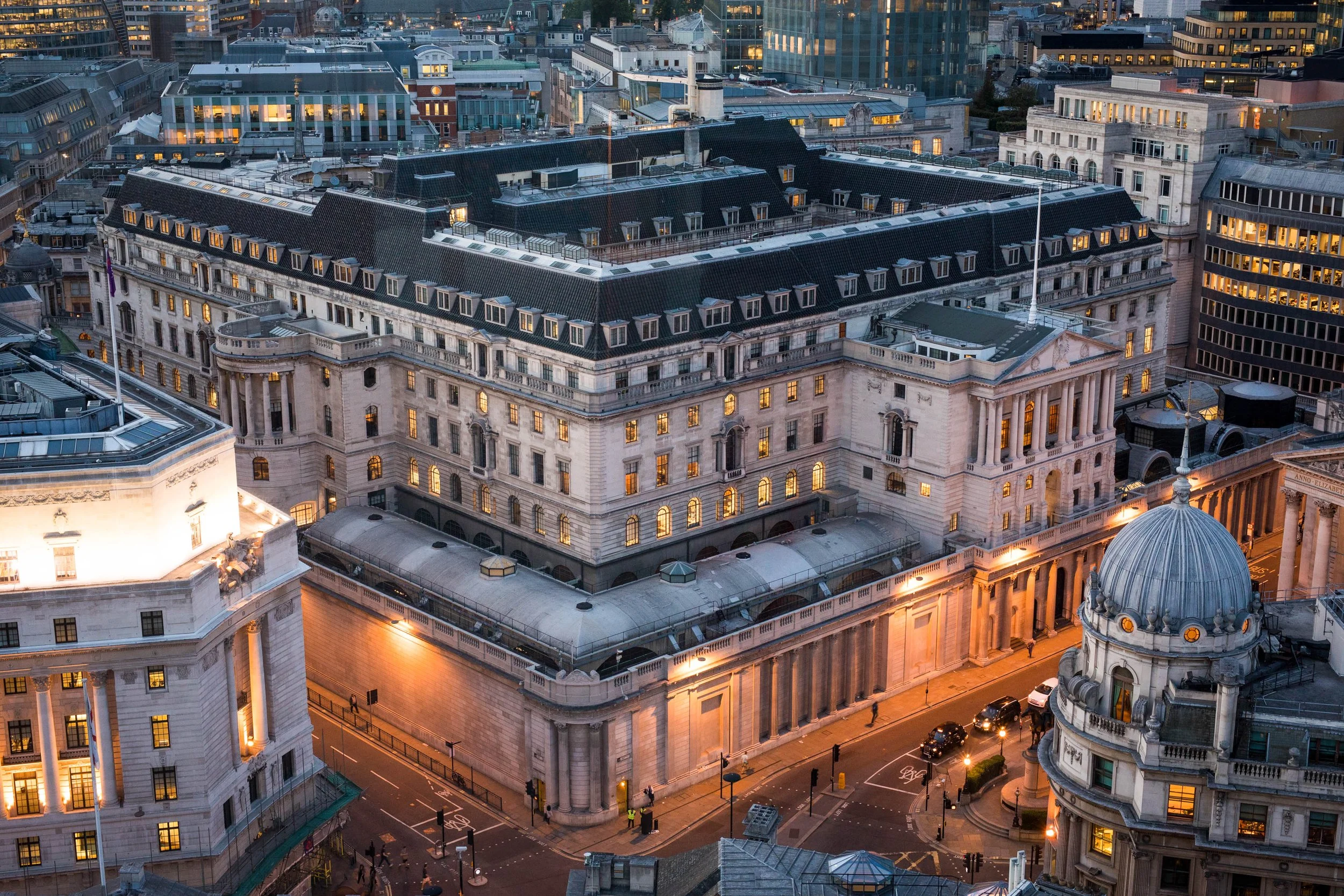 An aerial view of a historic building in a bustling city at dusk, with other modern buildings surrounding it, illuminated by streetlights and interior lighting.