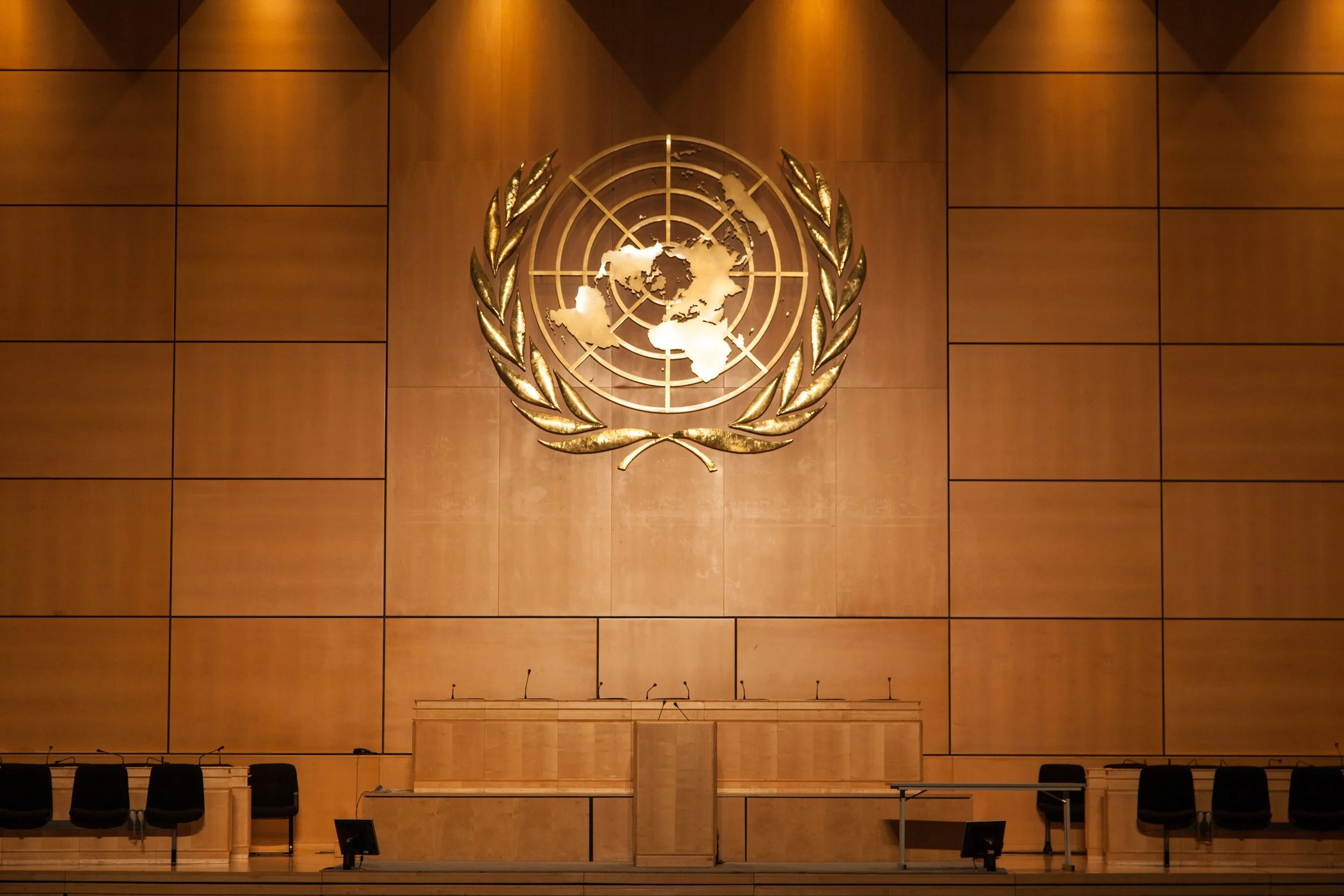 United Nations emblem on a large wooden wall in a conference room, with chairs and microphones set up in front.