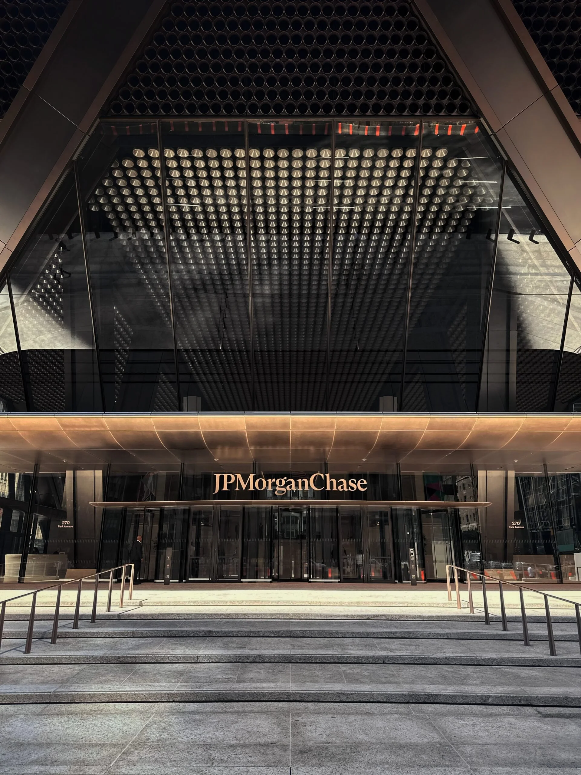 The entrance of a modern office building with glass doors and a sign that reads "JPMorgan Chase." The building has a distinctive geometric design with a large, triangular overhang and reflective glass facade.