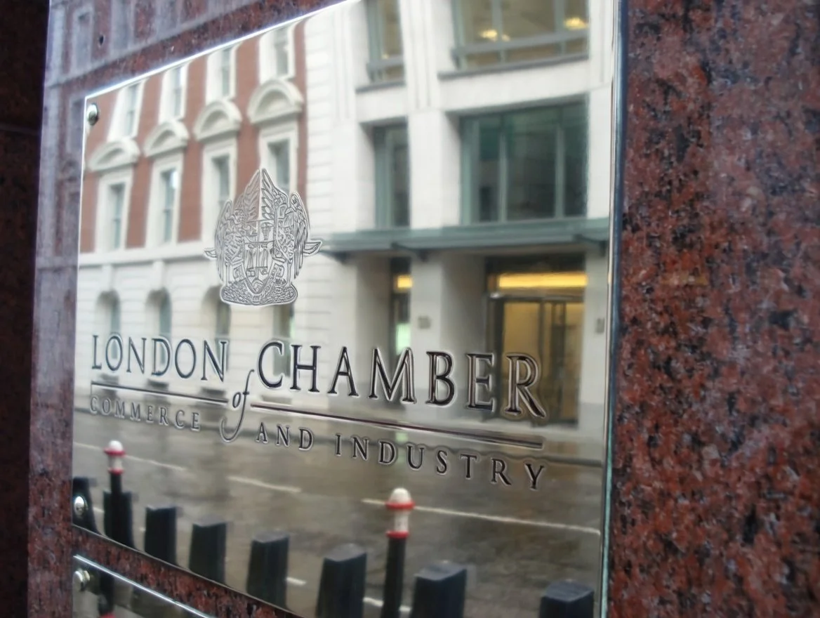 Metal sign with the London Chamber of Commerce and Industry emblem, reflecting a city street with buildings and bollards in the background.