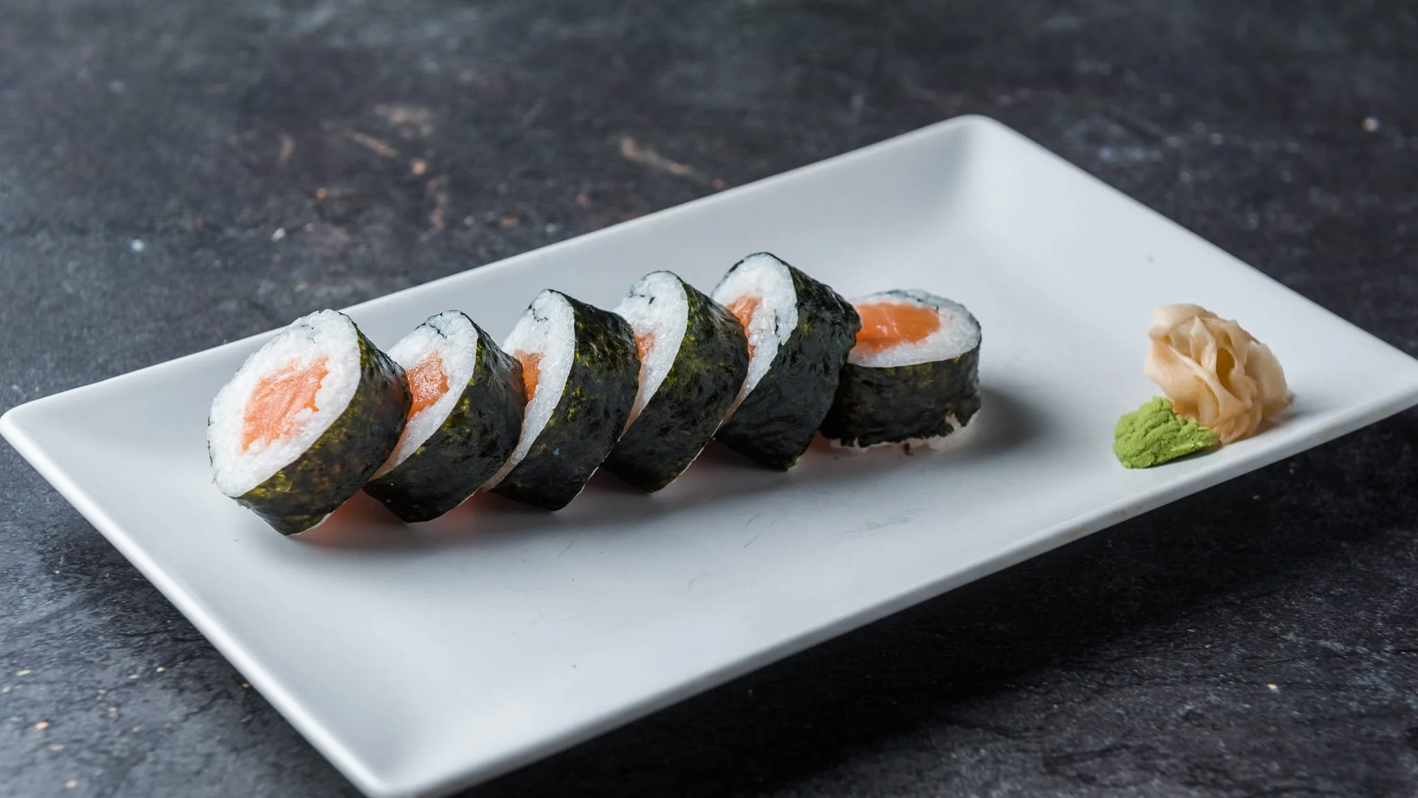 Six sushi rolls with salmon and rice, served on a rectangular white plate with wasabi and pickled ginger garnishes.