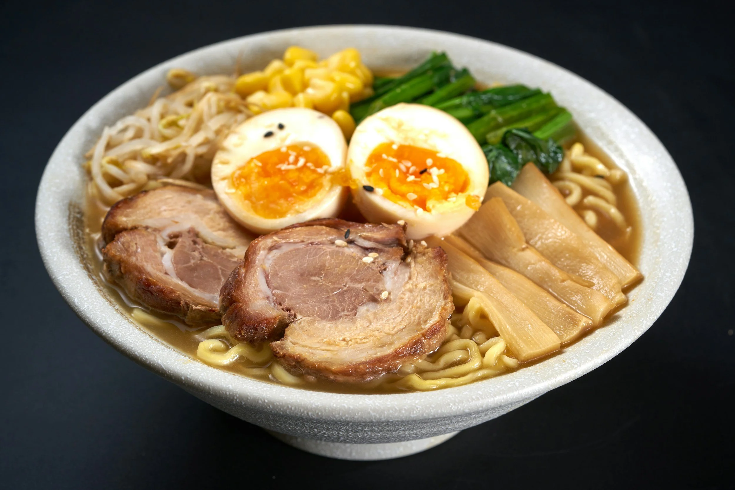 A bowl of ramen noodles with sliced pork, soft boiled egg halves, bamboo shoots, green vegetables, corn, and bean sprouts.