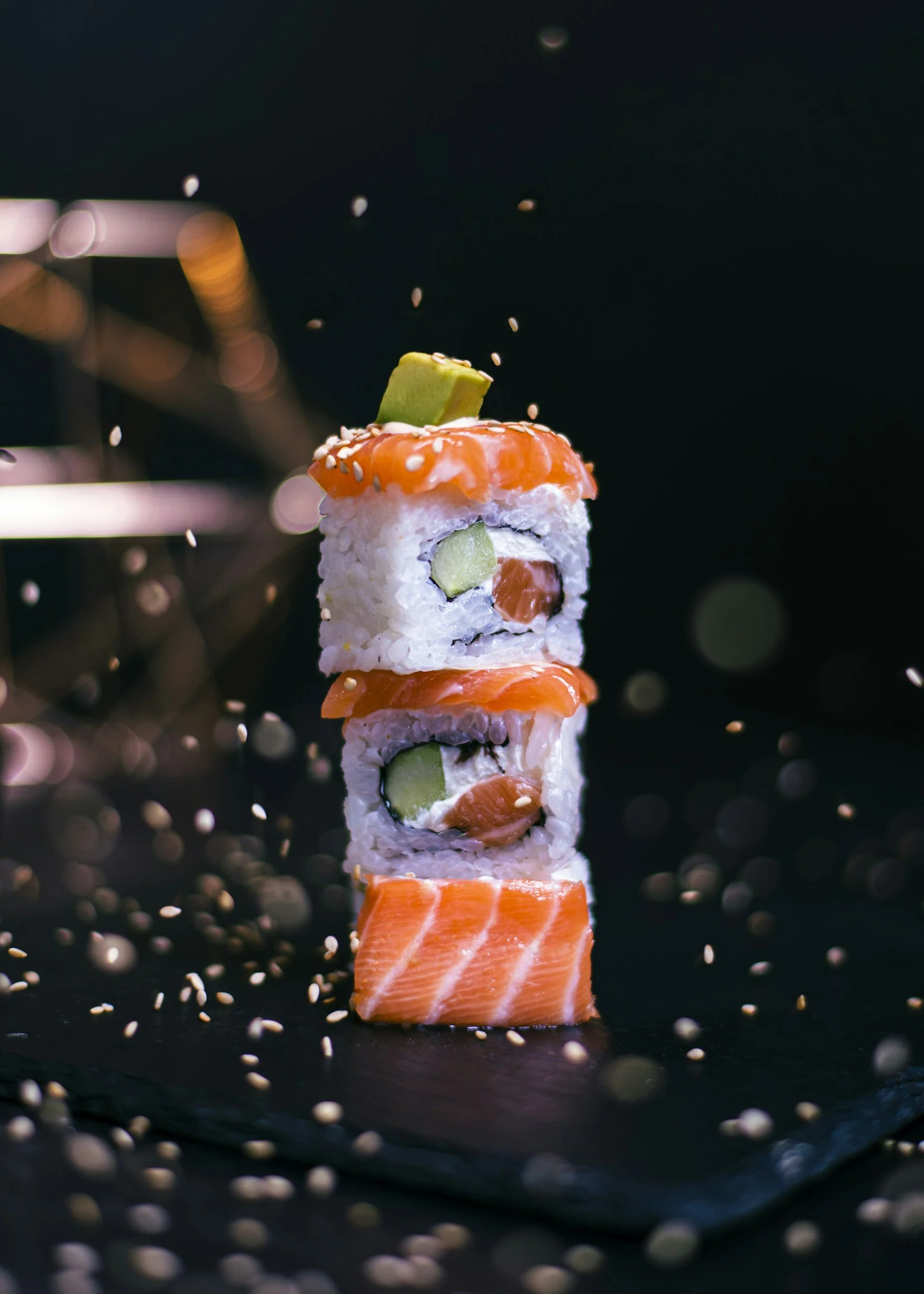 Sushi rolls stacked vertically with salmon, cucumber, and avocado, garnished with sesame seeds, on a black slate surface with scattered sesame seeds, and a dark background.