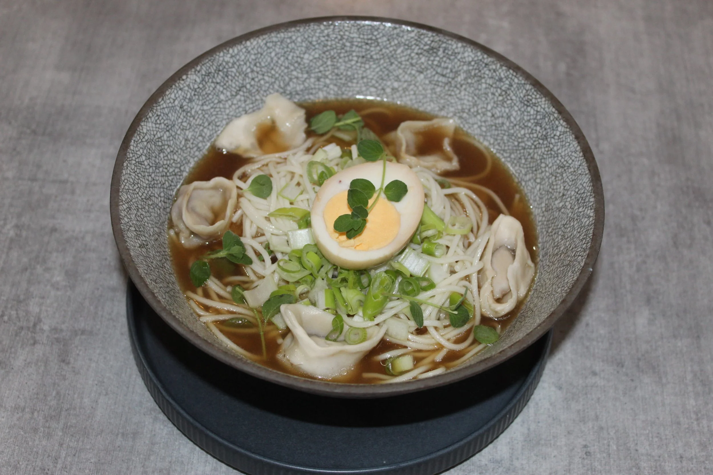 A bowl of ramen noodles with sliced boiled egg, chopped green onions, cilantro, and dumplings in broth.