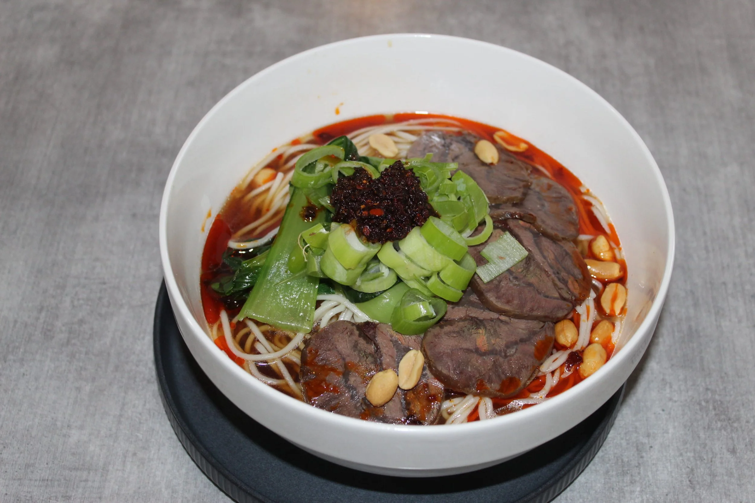 A bowl of Vietnamese pho with sliced beef, green onions, peanuts, and chili paste in broth.