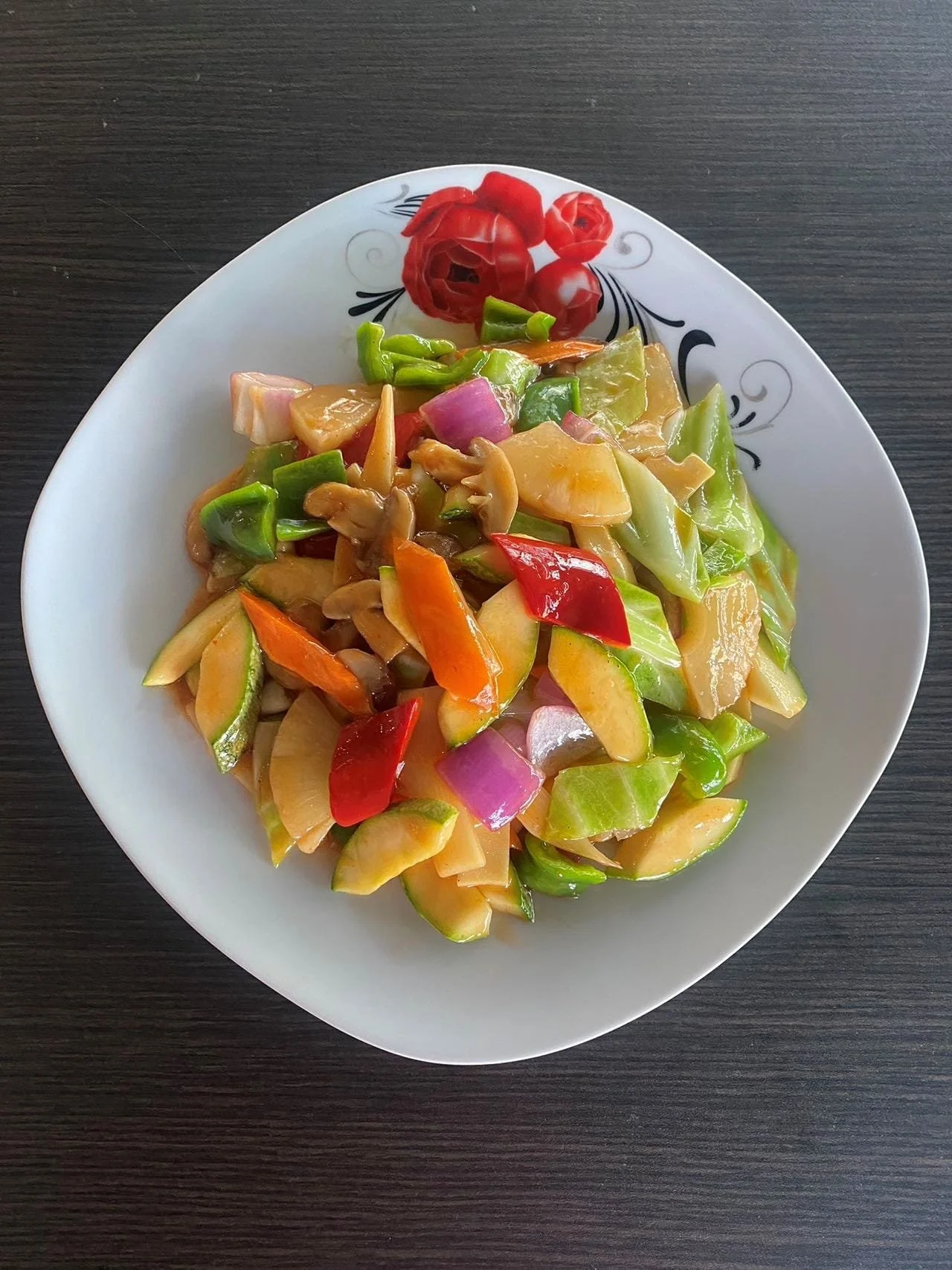 A white plate with a floral design on the rim, filled with a colorful mixed vegetable stir-fry on a dark wooden table.