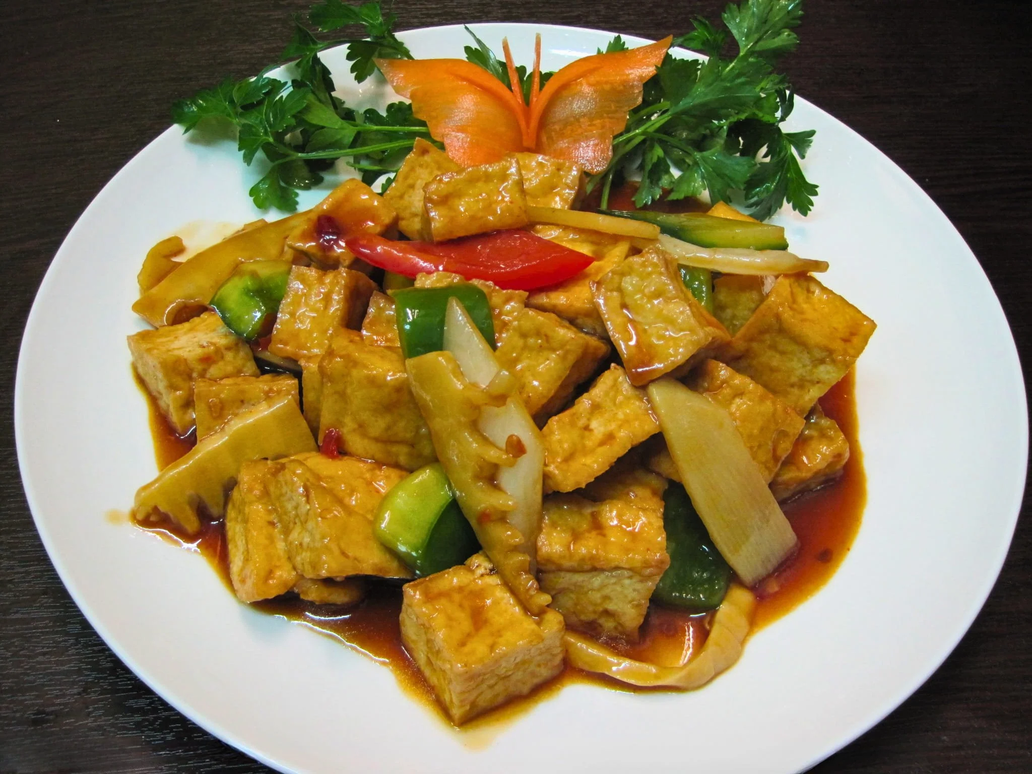 A white plate of stir-fried tofu with vegetables including bell peppers, onions, and zucchini, garnished with carved carrot and green herbs.