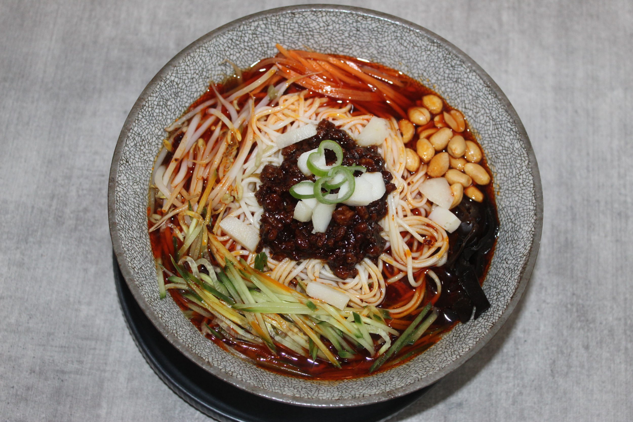 Bowl of Asian noodle soup with bean sprouts, ground meat, cucumbers, peanuts, and chili oil.