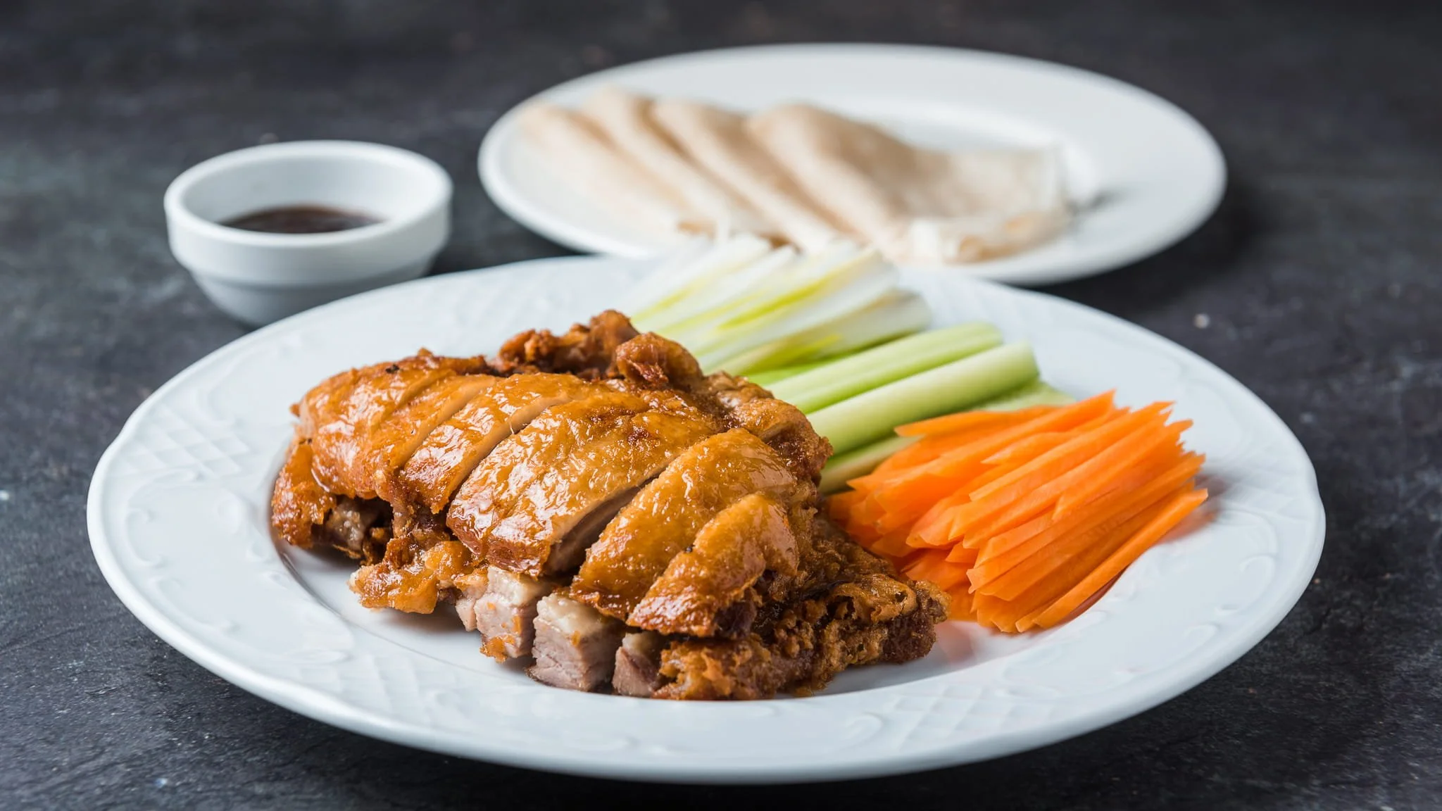 Plate of sliced roasted duck meat with fresh sliced vegetables including carrots, cucumbers, and white onions, with a small bowl of dipping sauce and a small plate of pancake in the background.