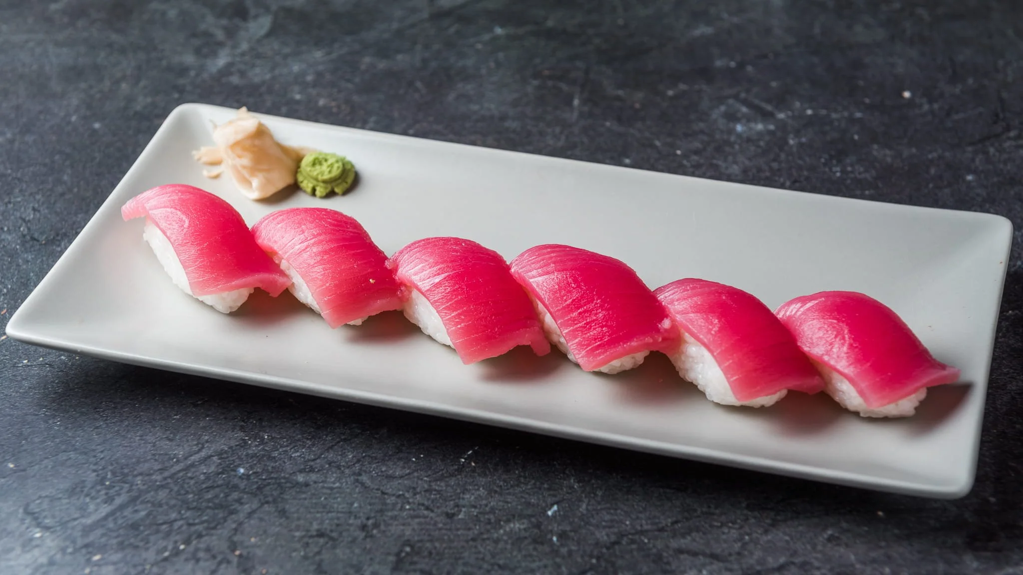 Sliced pink tuna nigiri sushi arranged on a white rectangular plate with wasabi and ginger on a dark surface.